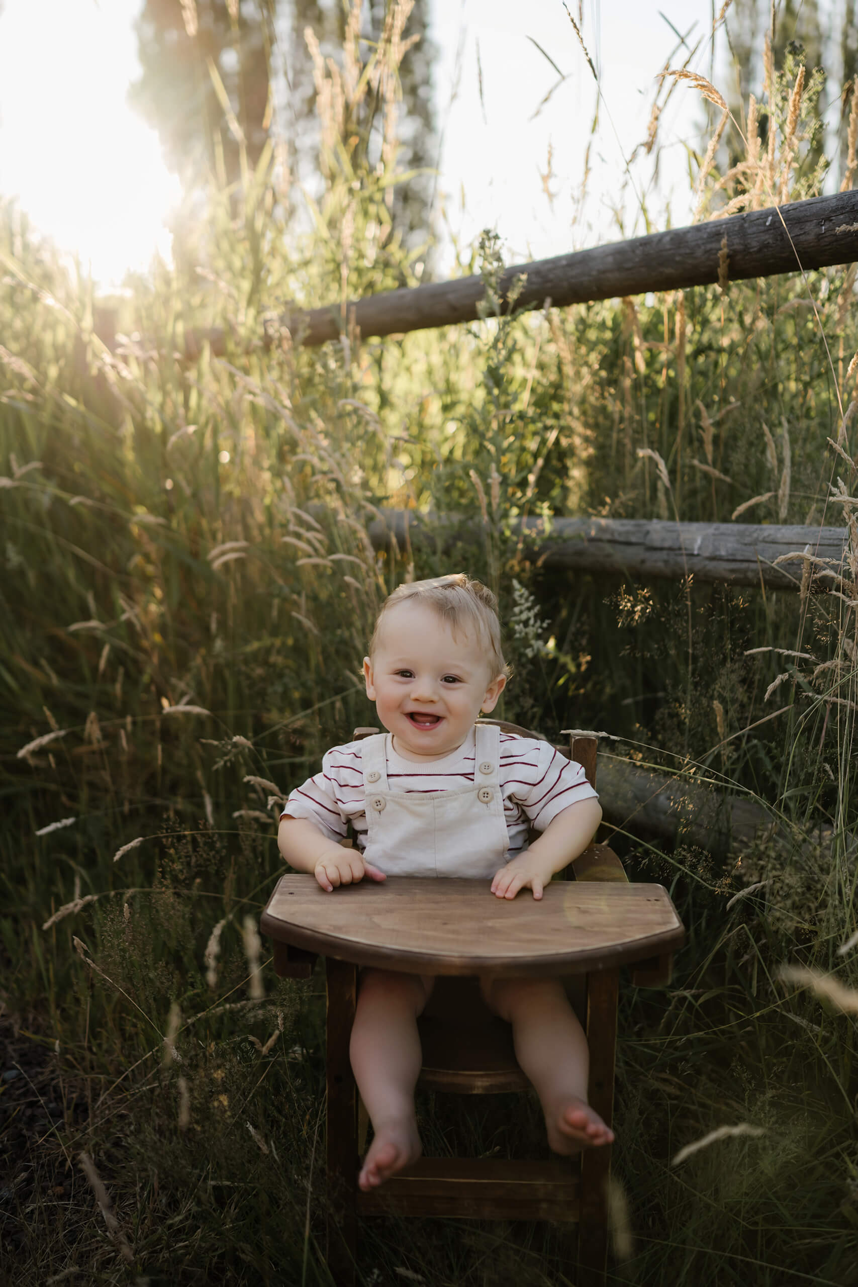 Smiling baby sits in a vintage wooden high-chair desk amid golden meadow grass and a sunlit split-rail fence.