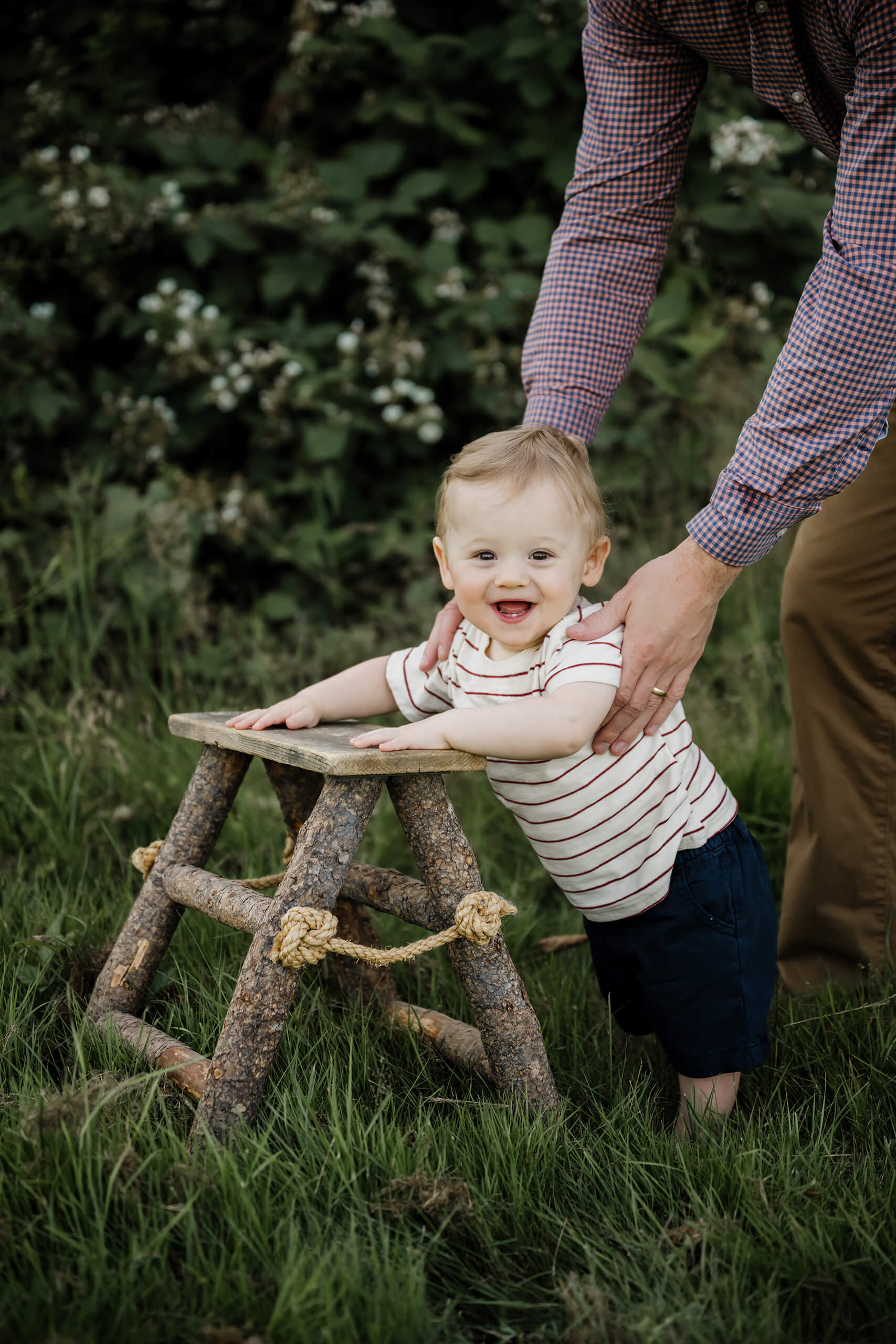 Dad steadies a grinning baby who’s standing and leaning on a rustic twig stool in the green grass.