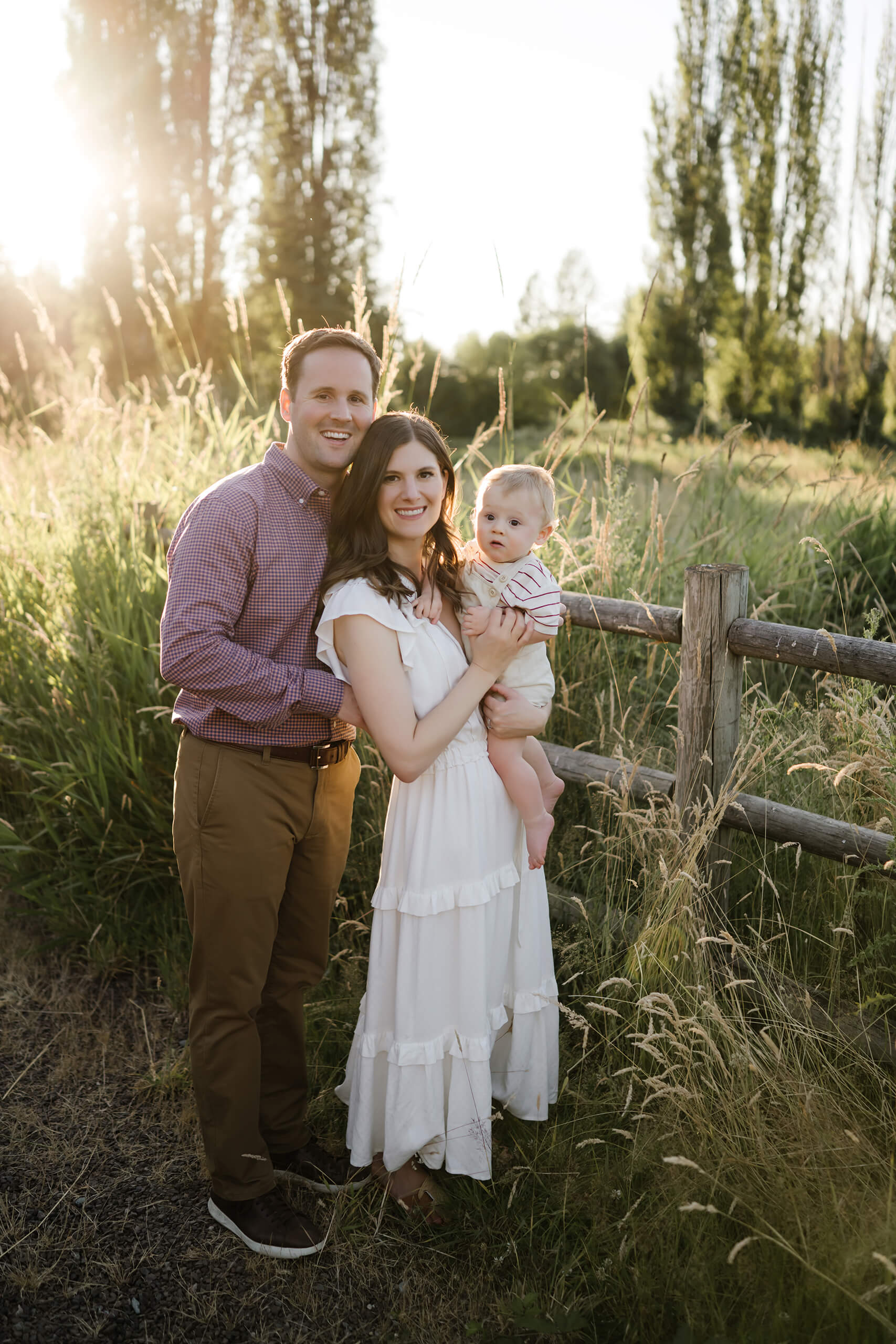 Back-lit family portrait with mom in a white dress holding baby by a wooden fence as dad hugs them, tall meadow grasses glowing in evening light.