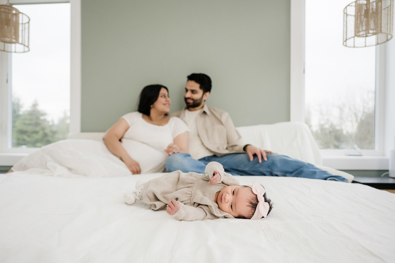 Newborn baby girl with a pink bow headband lying on the bed in front of her smiling parents during an in-home lifestyle photography session in Seattle.