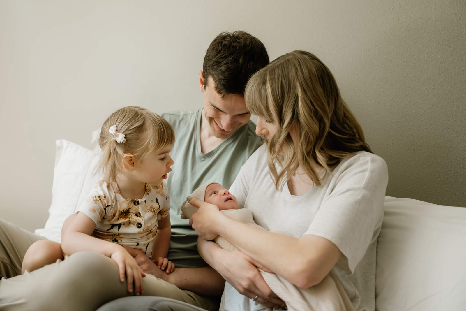 Family of four snuggling on the bed during an in-home newborn session in Seattle, with mom holding the baby, dad smiling, and big sister looking curiously at her new sibling