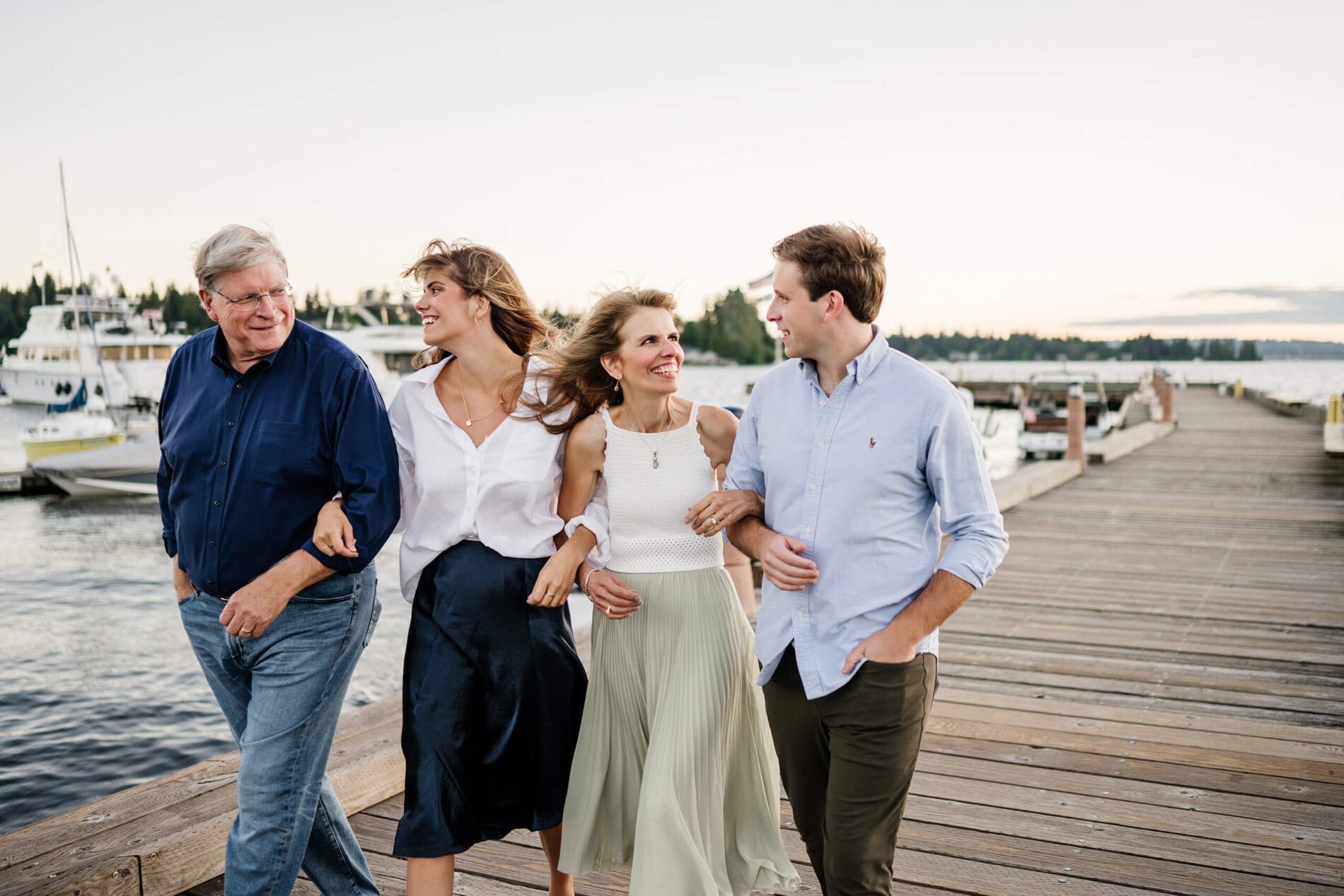 Family of four laughing and walking arm-in-arm down the Carillon Point dock at golden hour, yachts and Lake Washington behind them in Kirkland WA.