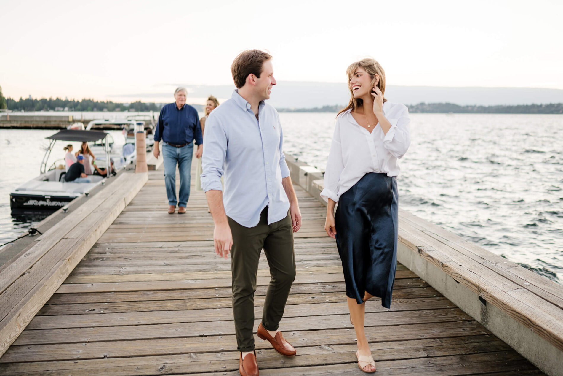 Adult brother and sister chatting and smiling on the wooden dock while parents follow behind, breezy summer evening at Carillon Point, Kirkland WA.