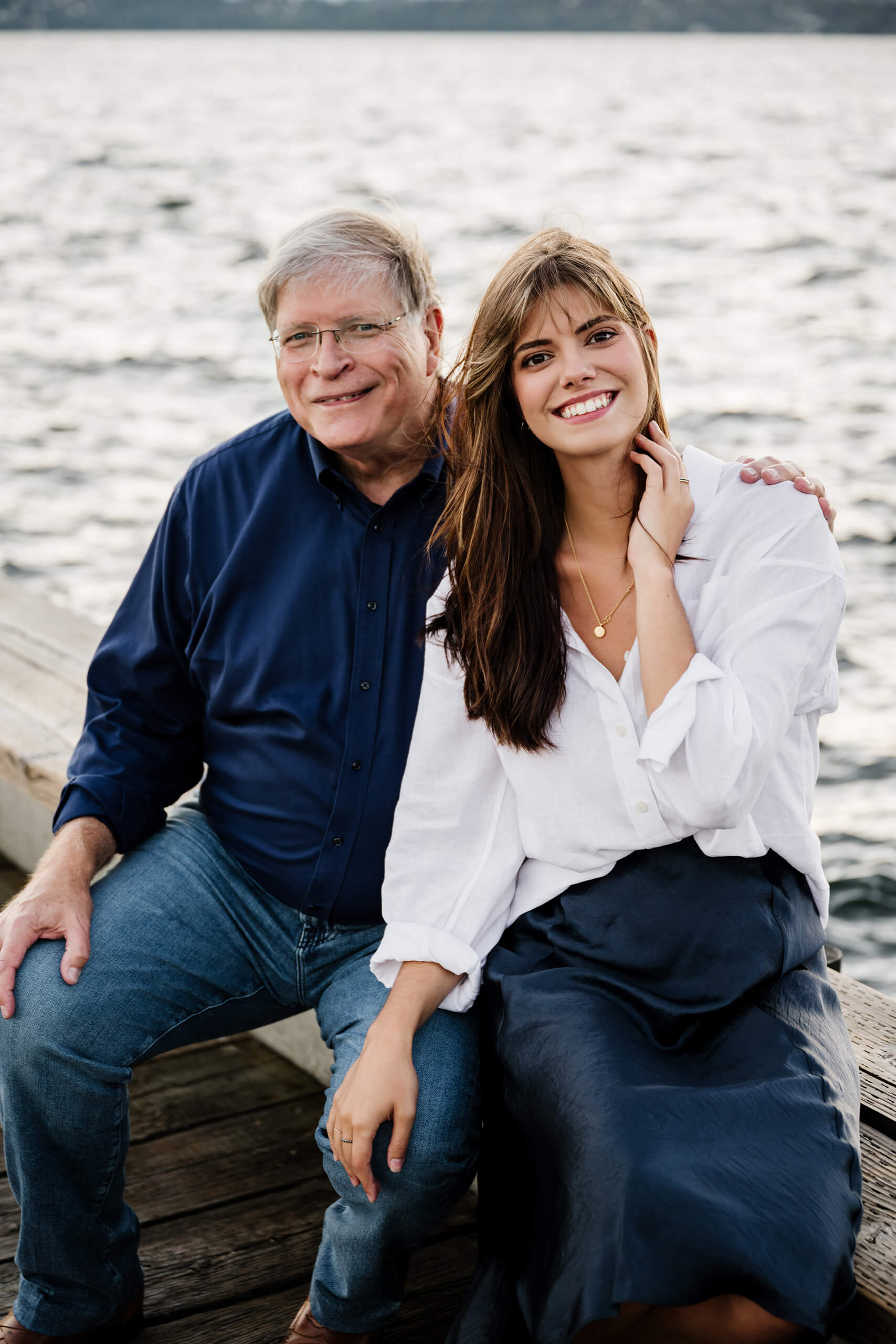 Father seated next to daughter, both smiling toward camera with rippling Lake Washington water as backdrop on the Carillon Point pier, Kirkland WA.