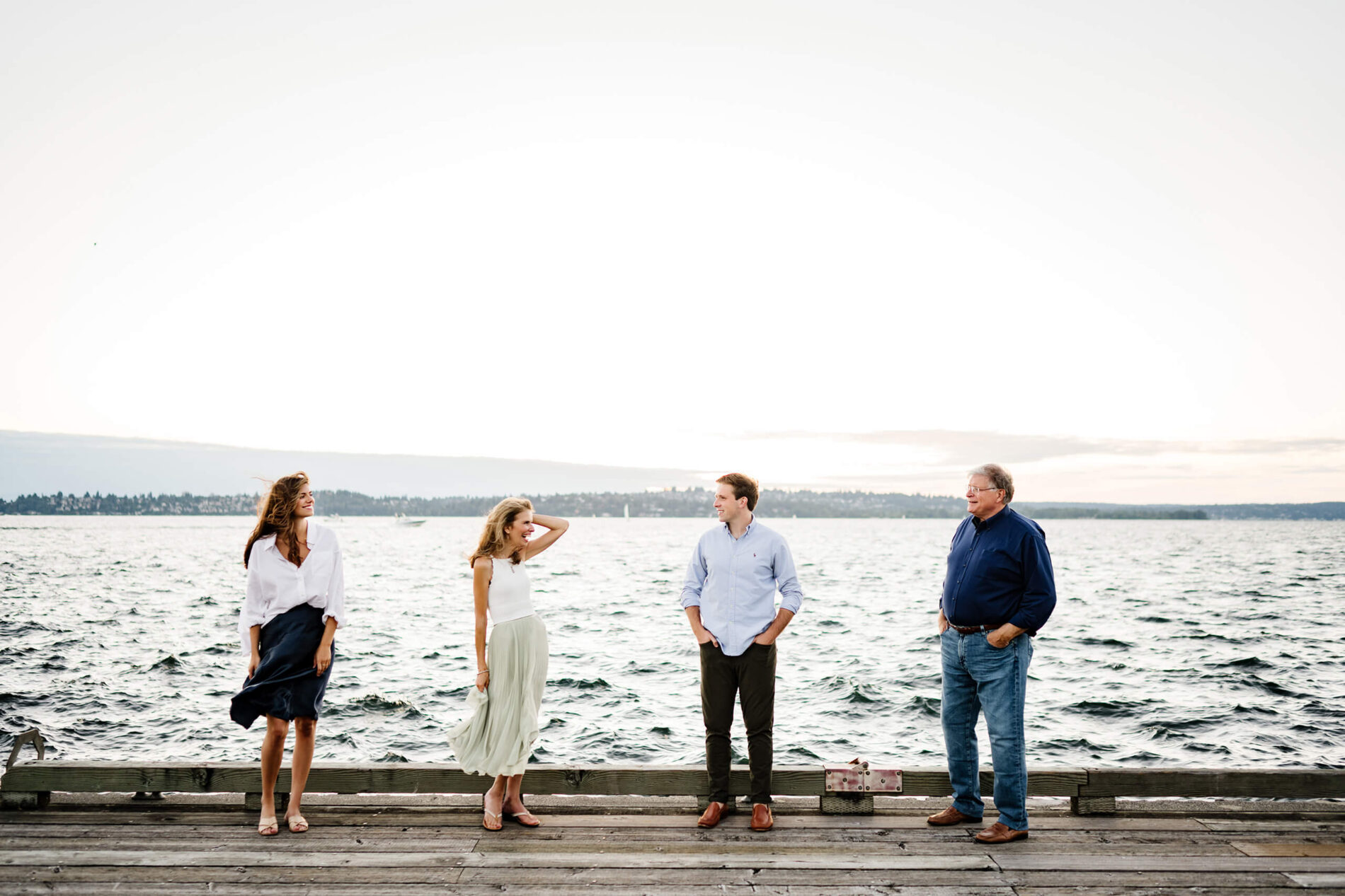 Windy sunset portrait of family spaced along dock edge, playful pose with Lake Washington vista at Carillon Point in Kirkland WA.