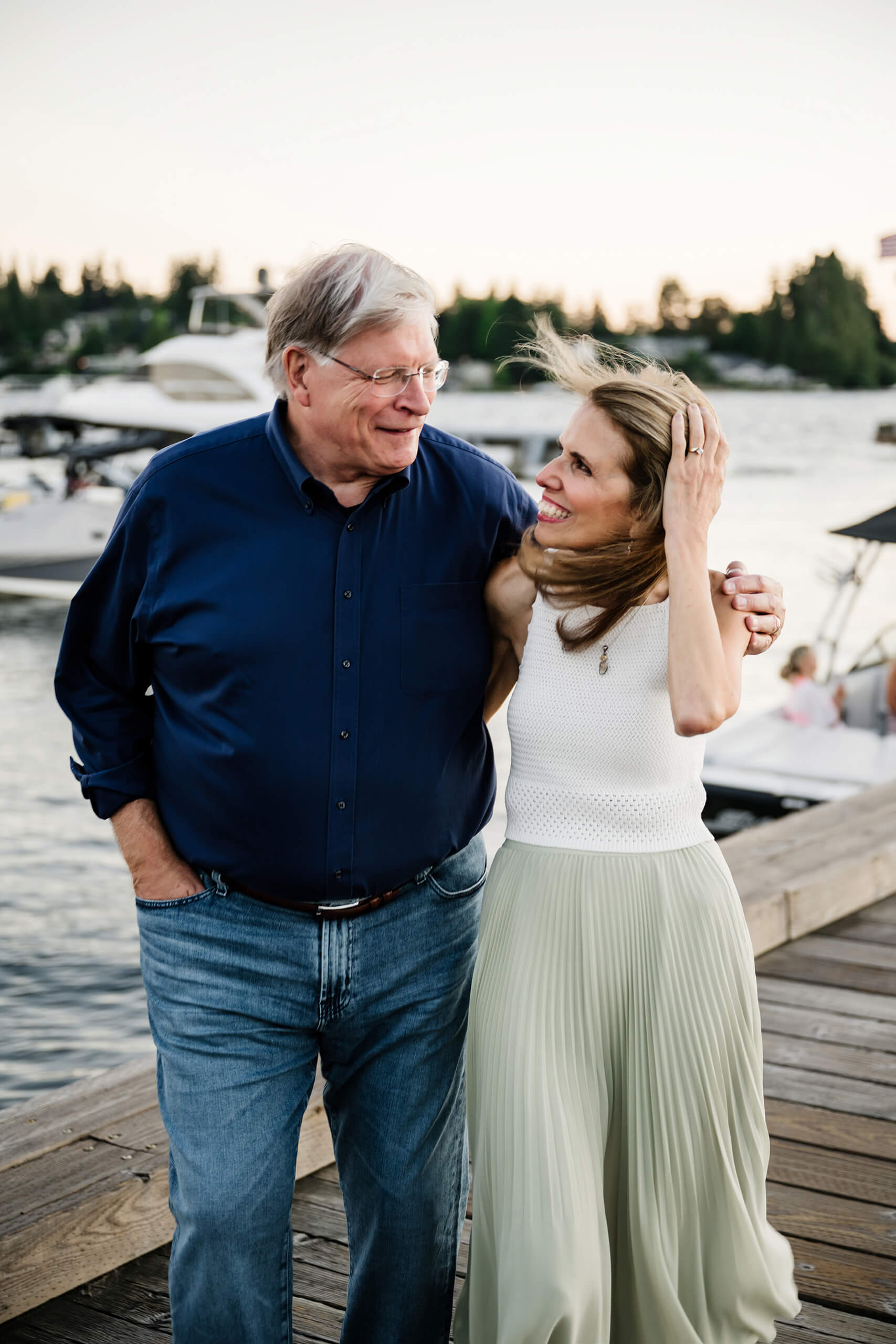 Husband and wife laughing arm-in-arm while strolling the Carillon Point boardwalk at sunset, boats softly blurred behind them in Kirkland WA.