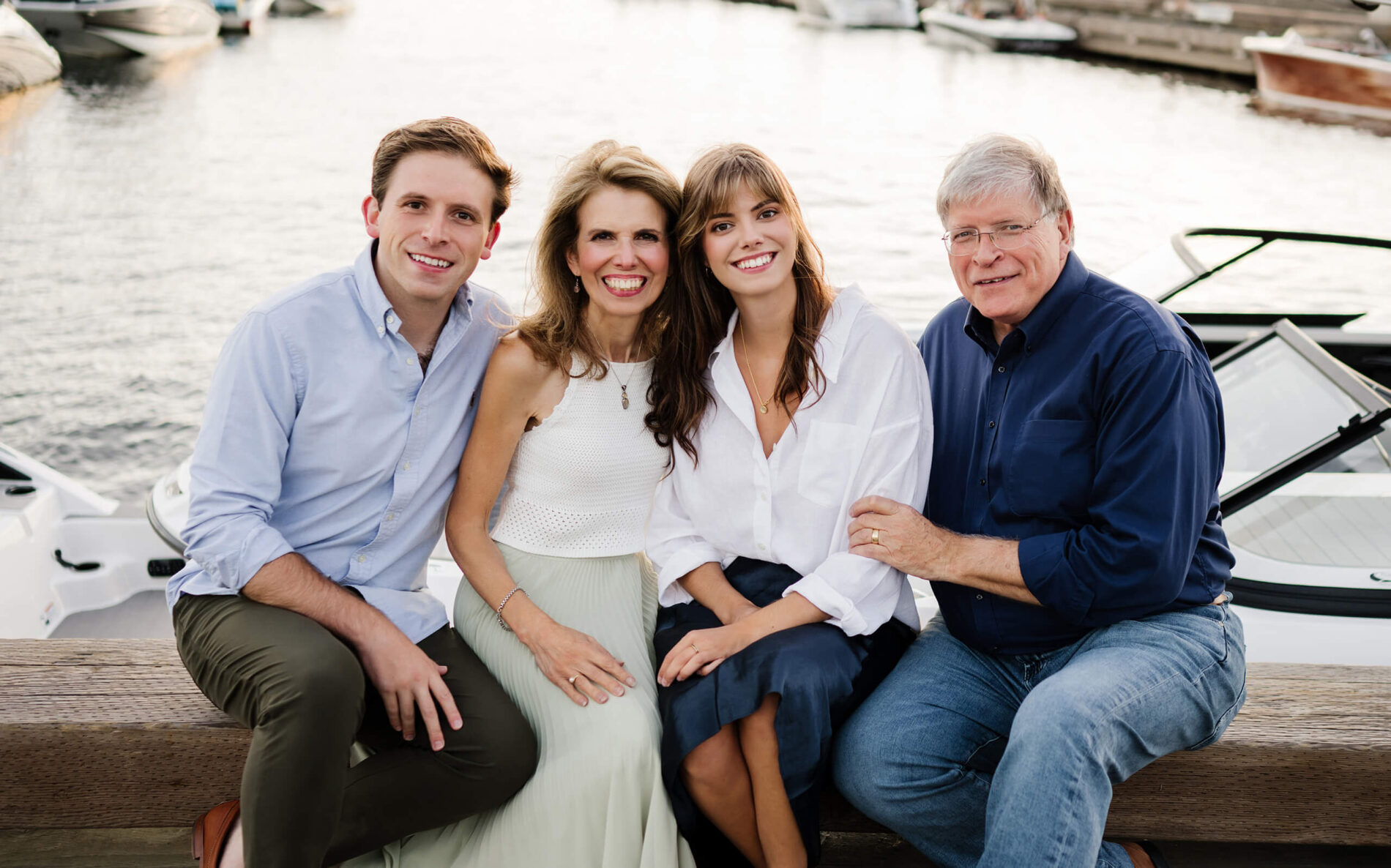Family of four seated side-by-side on a wooden bench beside a boat, smiling toward camera during a Carillon Point photo session in Kirkland WA.