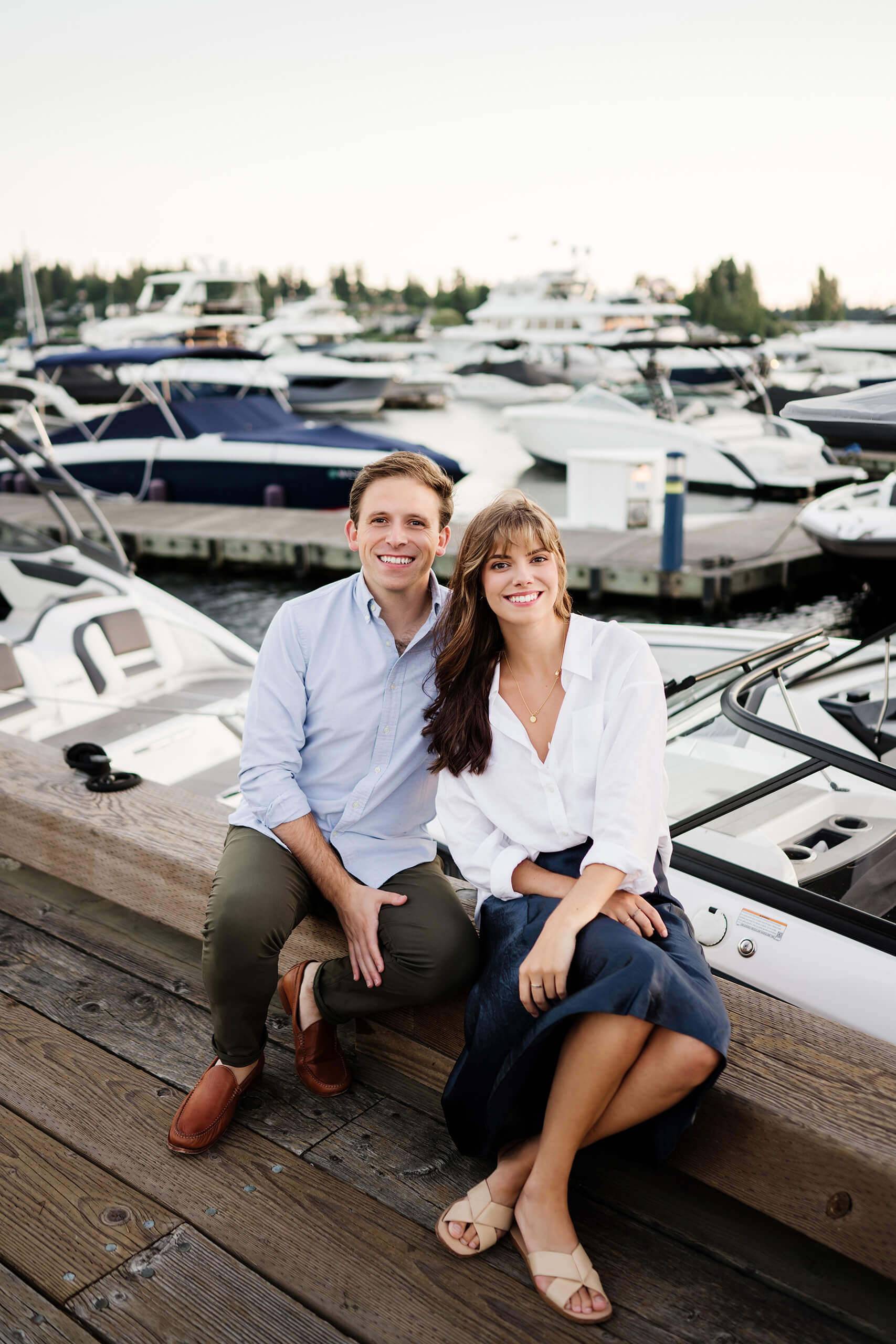 Adult brother and sister sitting together on the dock rail, rows of moored powerboats filling the Carillon Point marina behind them in Kirkland WA.