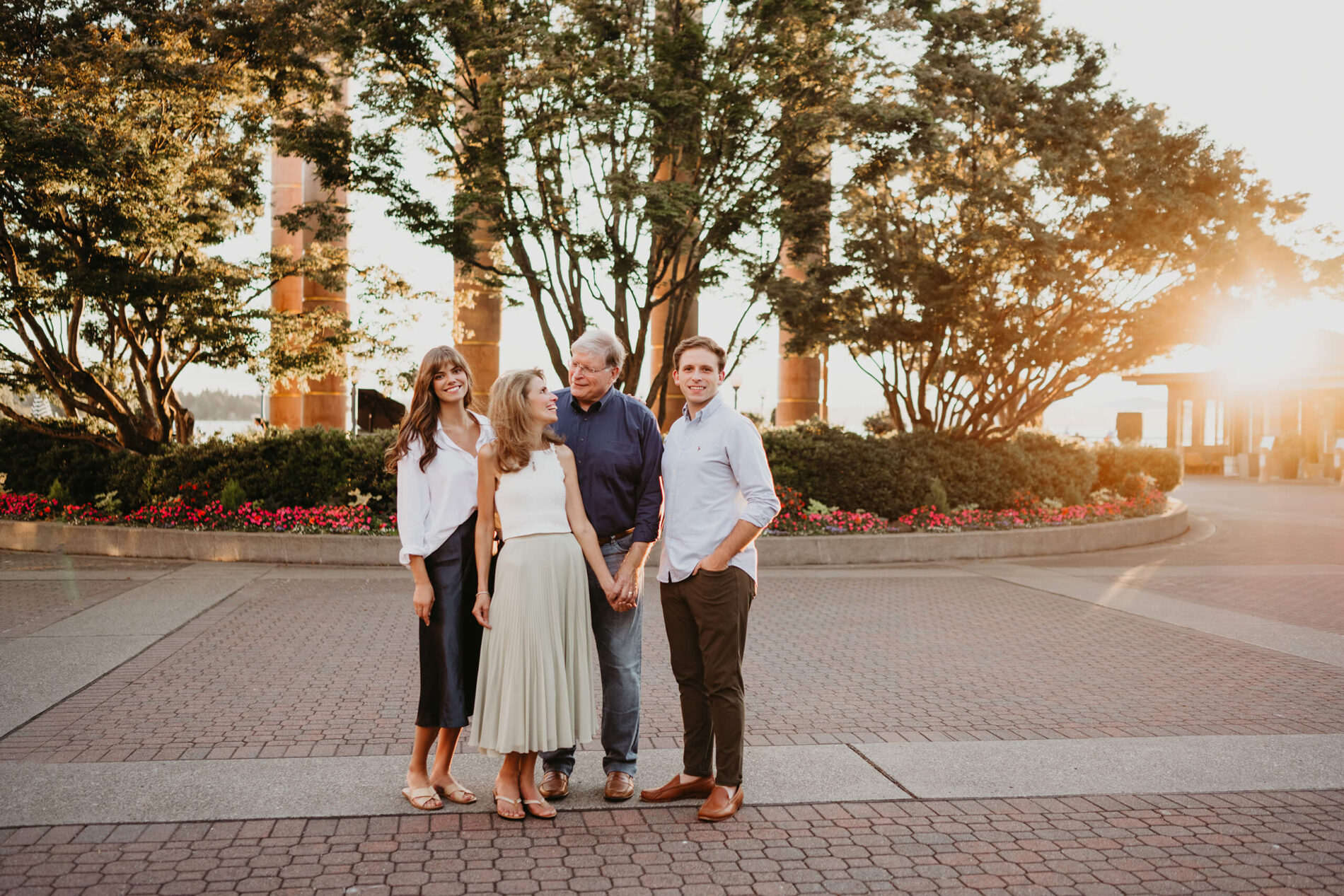 Family of four standing in the Carillon Point plaza at sunset, tree-lined promenade and art columns bathed in soft sun flare, Kirkland WA.