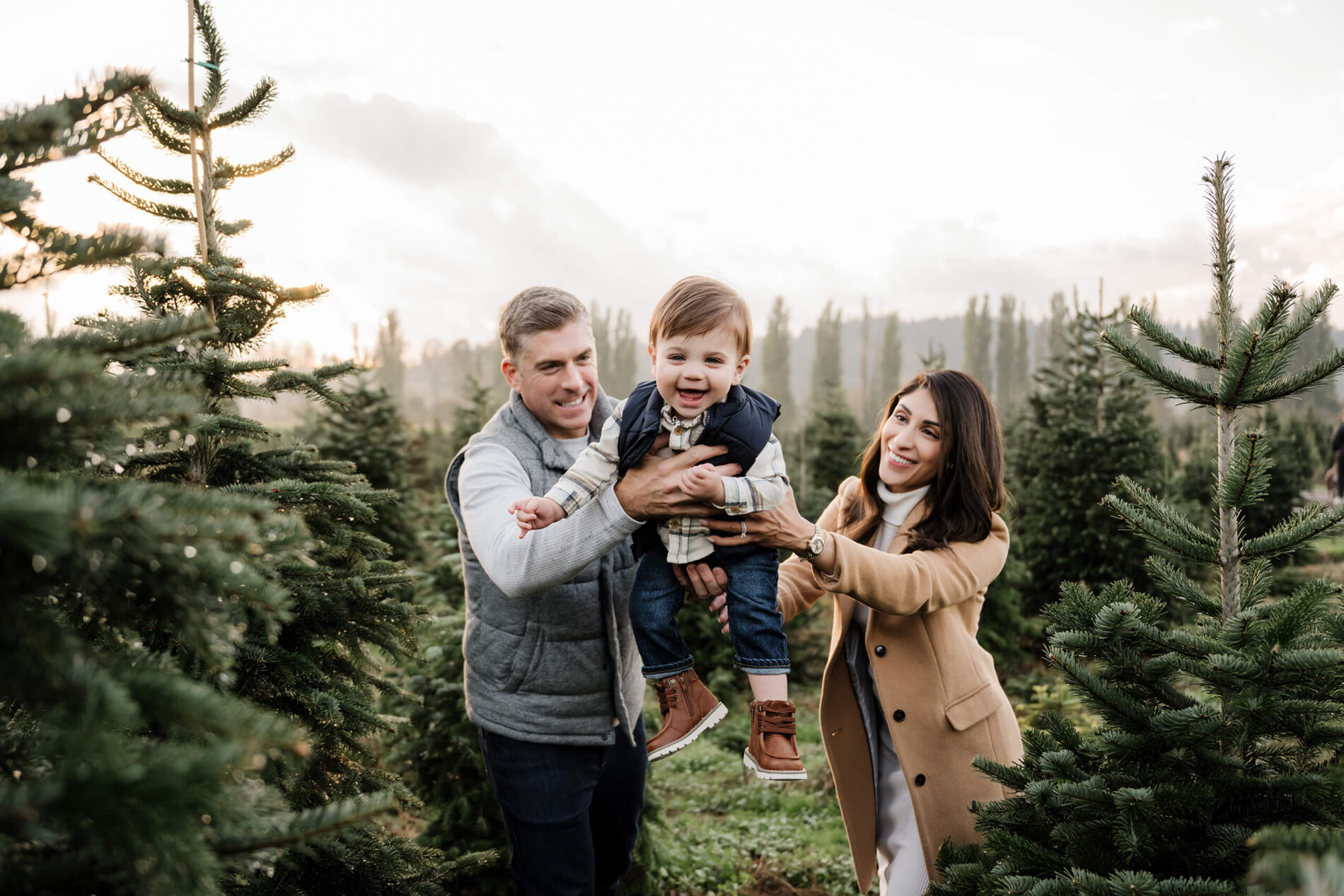 Parents lifting toddler at Christmas tree farm – Joyful Seattle Christmas tree farm family photo with young child laughing between mom and dad.