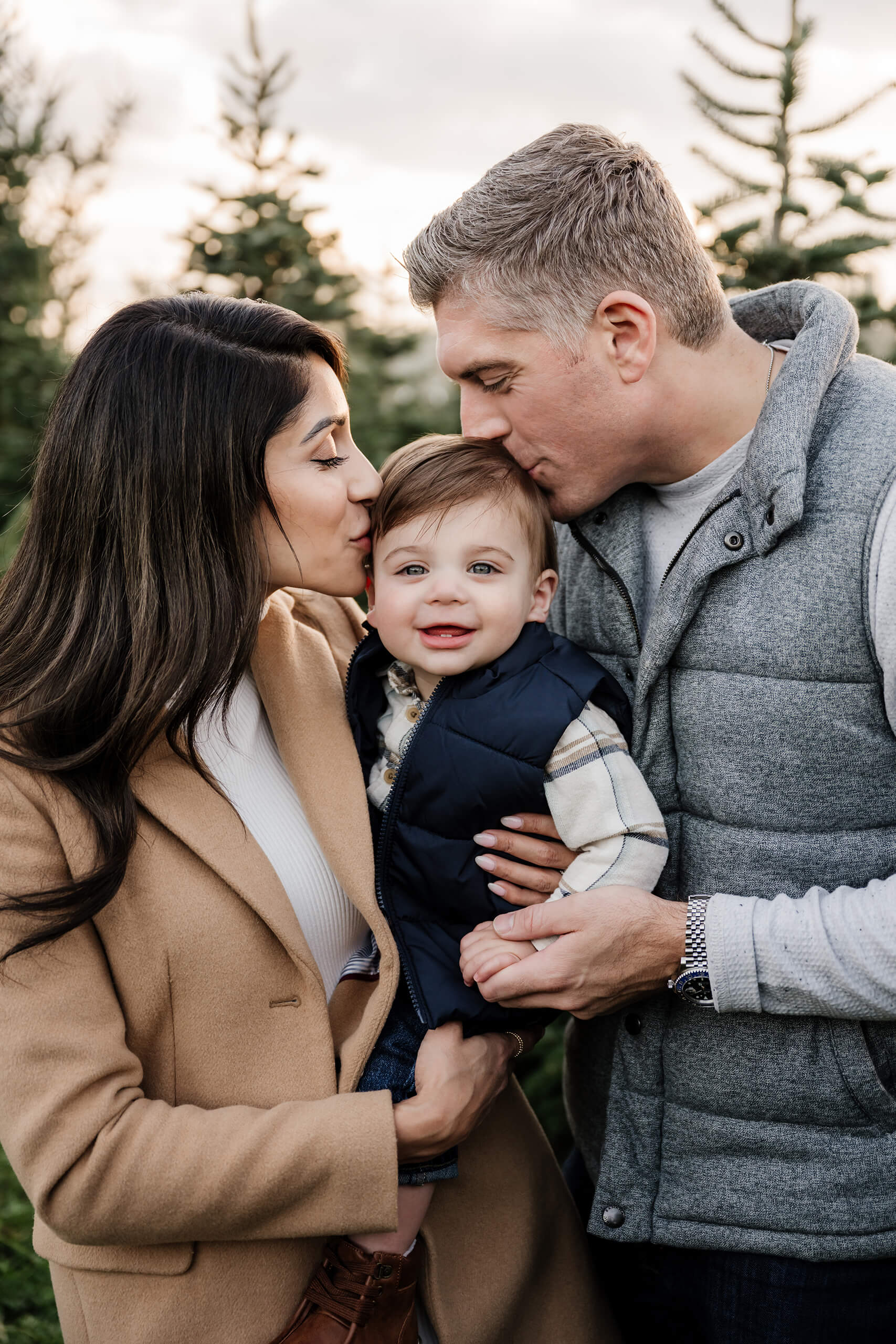Parents kissing toddler – Cozy family portrait during Seattle holiday mini session at tree farm.