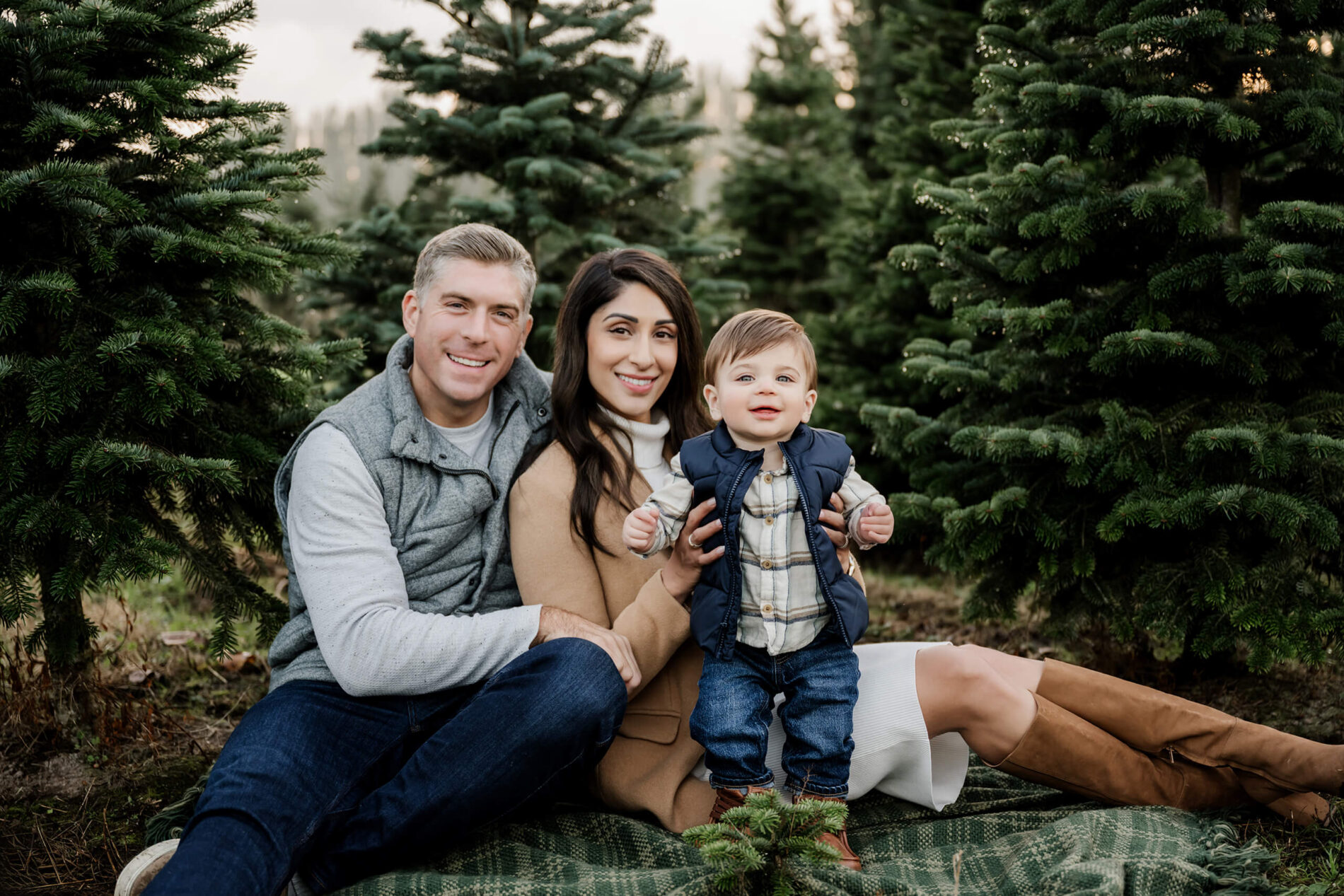 Family sitting on blanket – Seattle tree farm holiday photos featuring parents and toddler among evergreens.