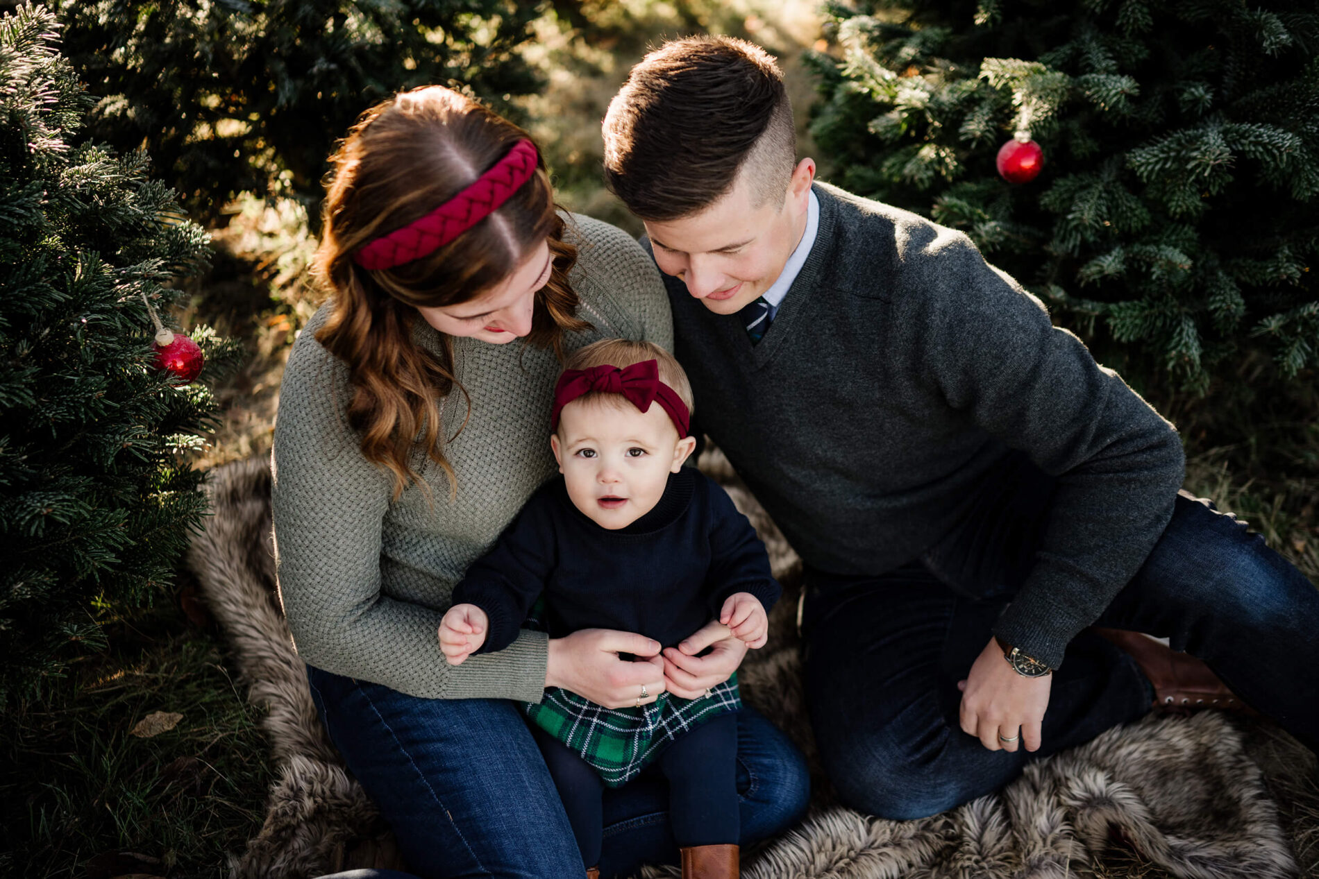 Parents sitting with toddler – Holiday mini session at Seattle Christmas tree farm with baby girl in plaid skirt and headband.