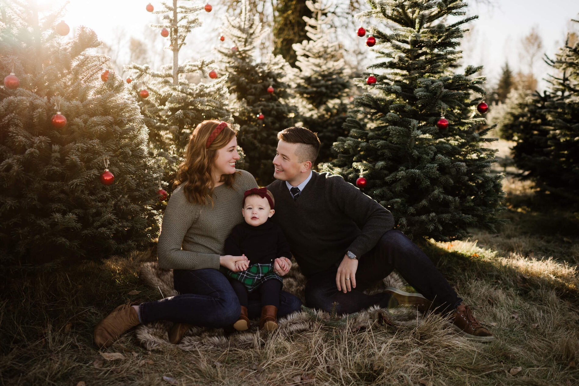 Family sitting on blanket – Seattle Christmas tree farm mini sessions with parents and toddler among decorated trees.