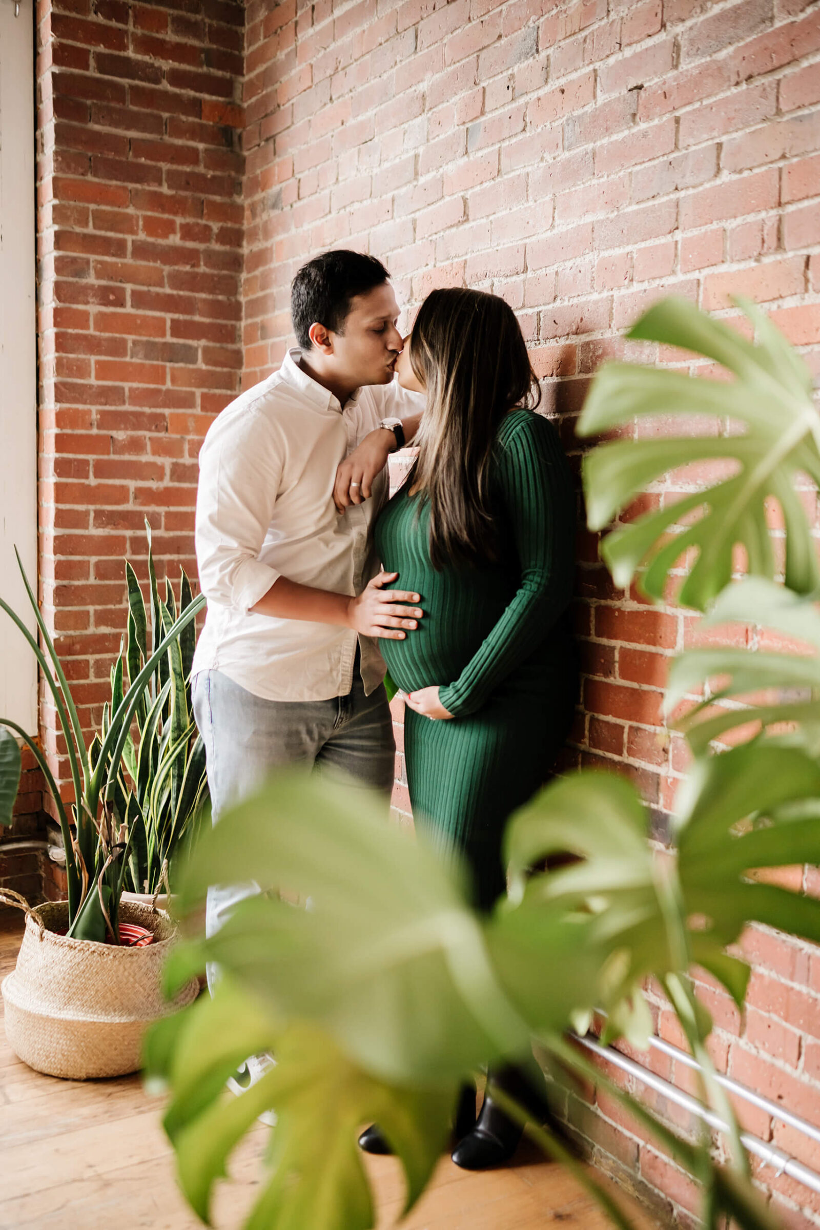 Expecting couple kissing by brick wall with green dress maternity outfit during Seattle studio session.
