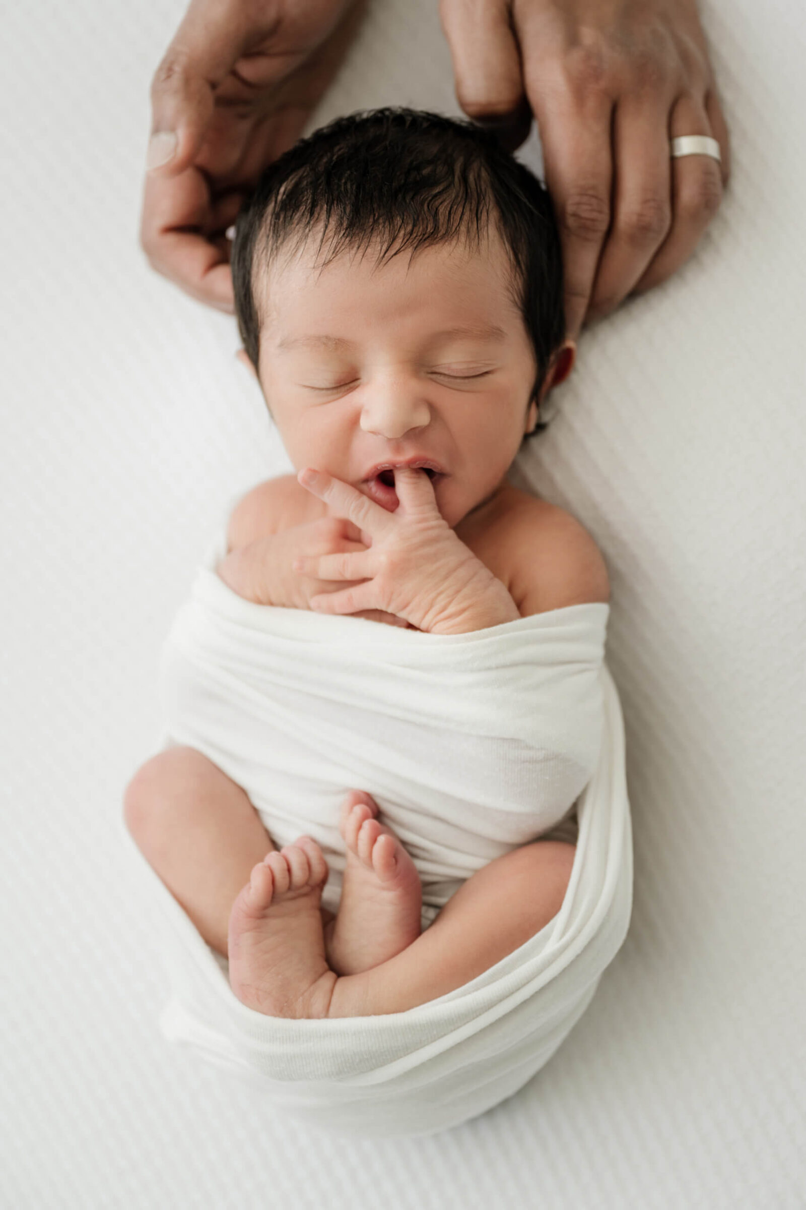 Baby swaddled in white wrap sucking on fingers during Seattle studio photo session.