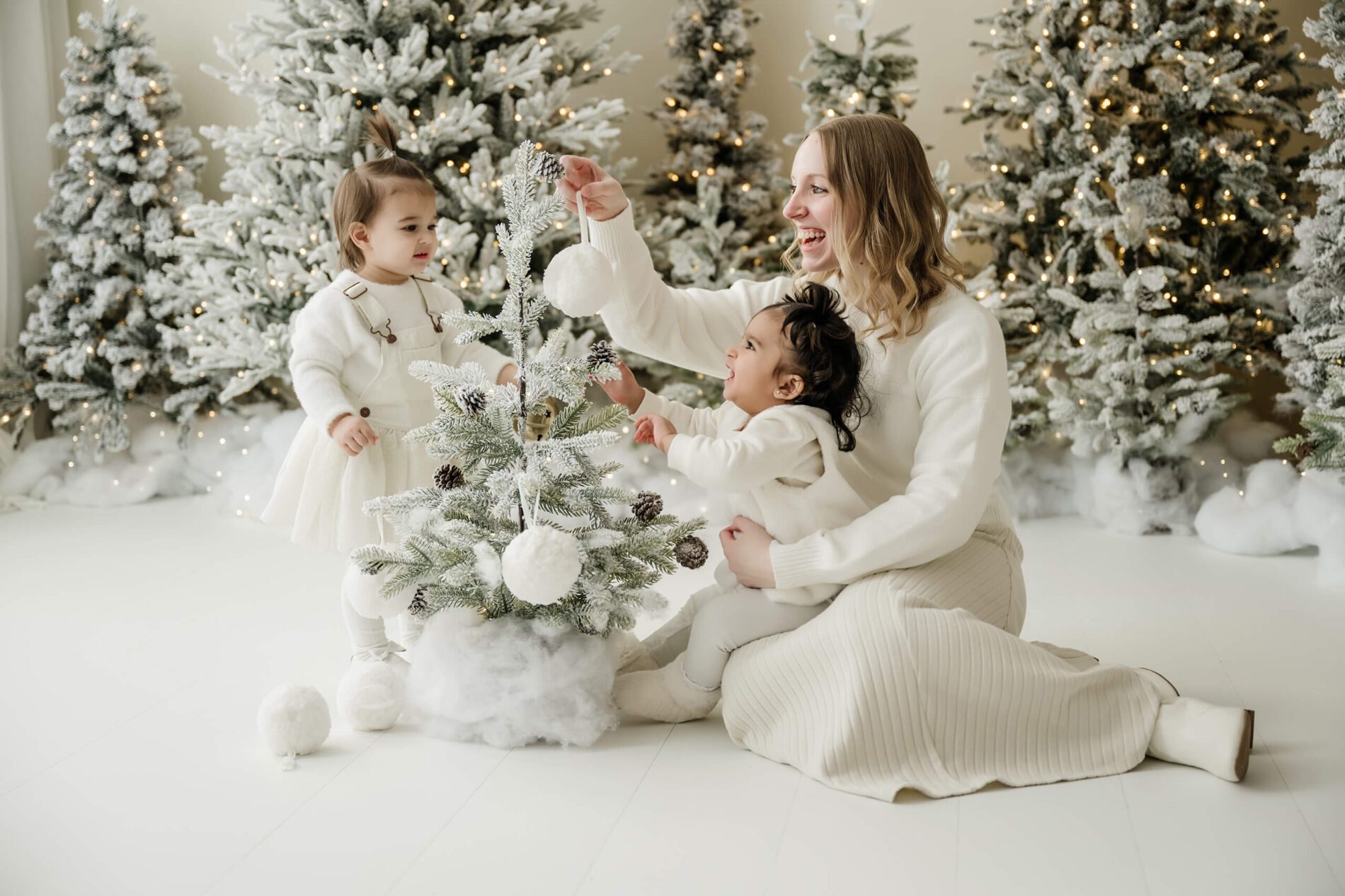 Mother decorating small Christmas tree with two toddlers during Seattle holiday studio photo session in Kirkland WA, surrounded by snow-flocked trees and twinkle lights.