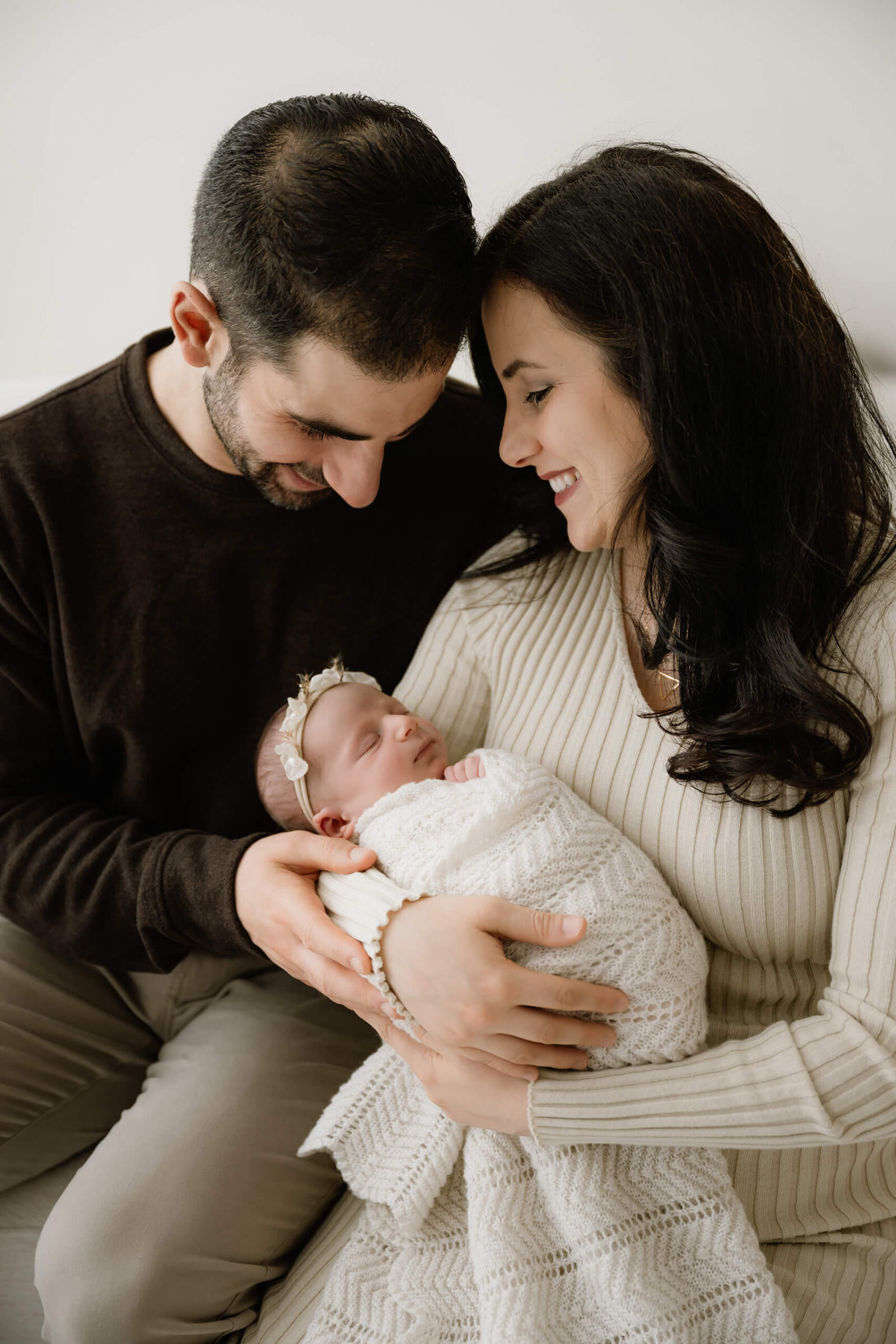 Mother and father gazing lovingly at their sleeping newborn baby wrapped in a soft white blanket during a cozy Kirkland studio newborn session by Lana Sky Photography.