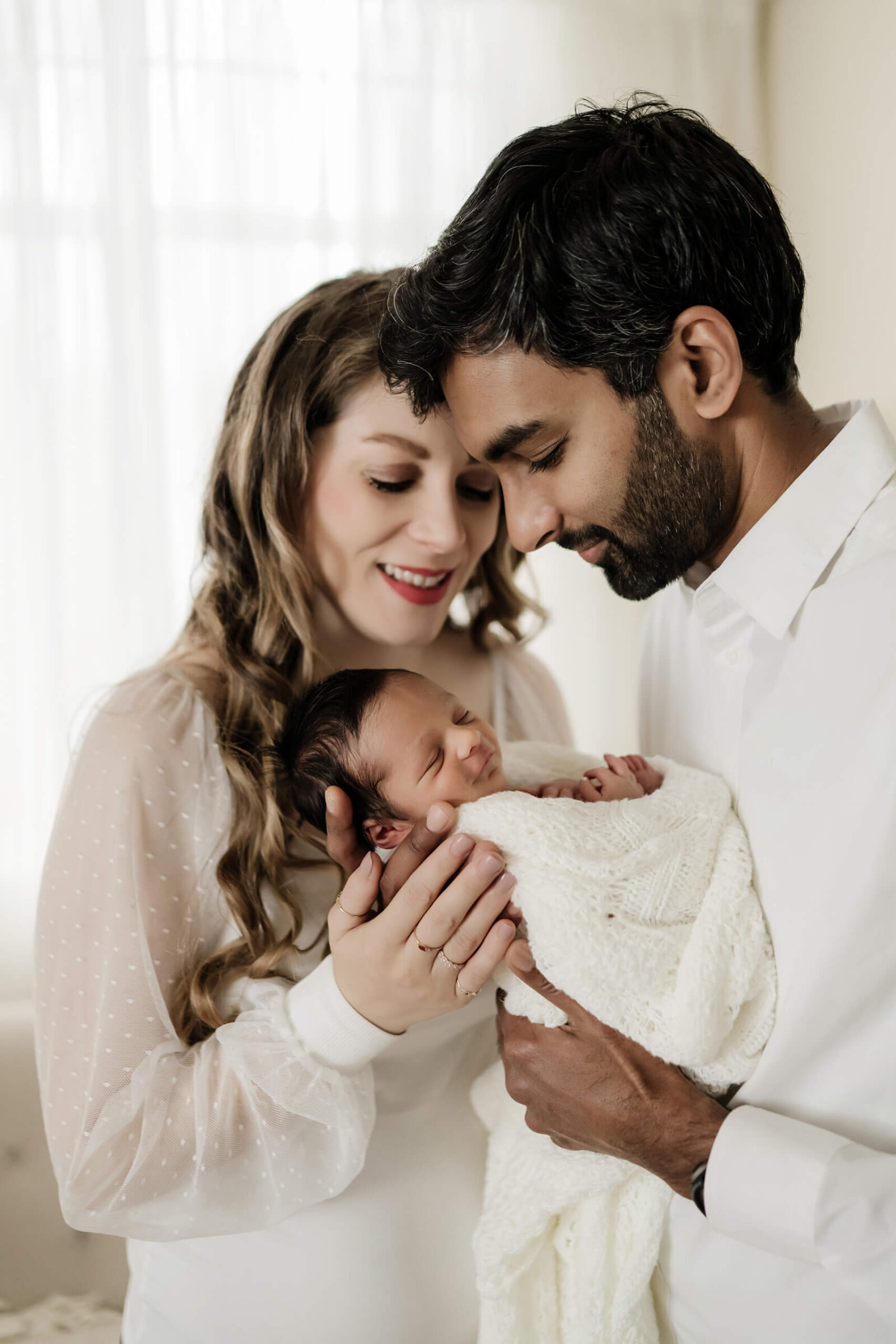 Parents gently cradling their sleeping newborn wrapped in white blanket during a bright and natural newborn photoshoot.