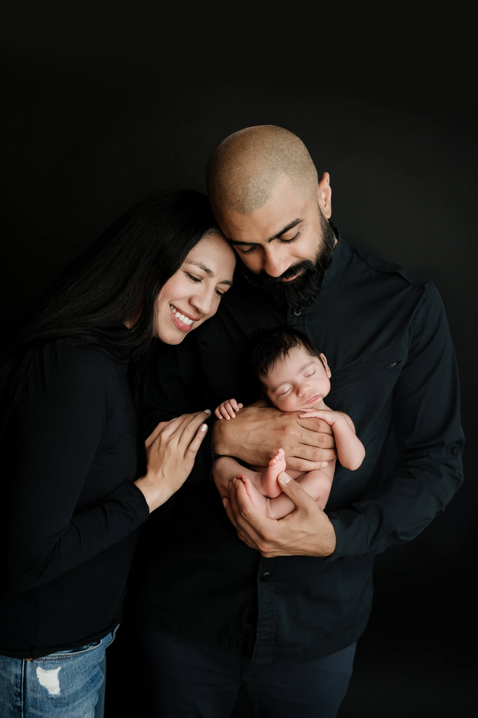 Parents dressed in black smiling down at their newborn baby during an intimate studio photo session for newborn.