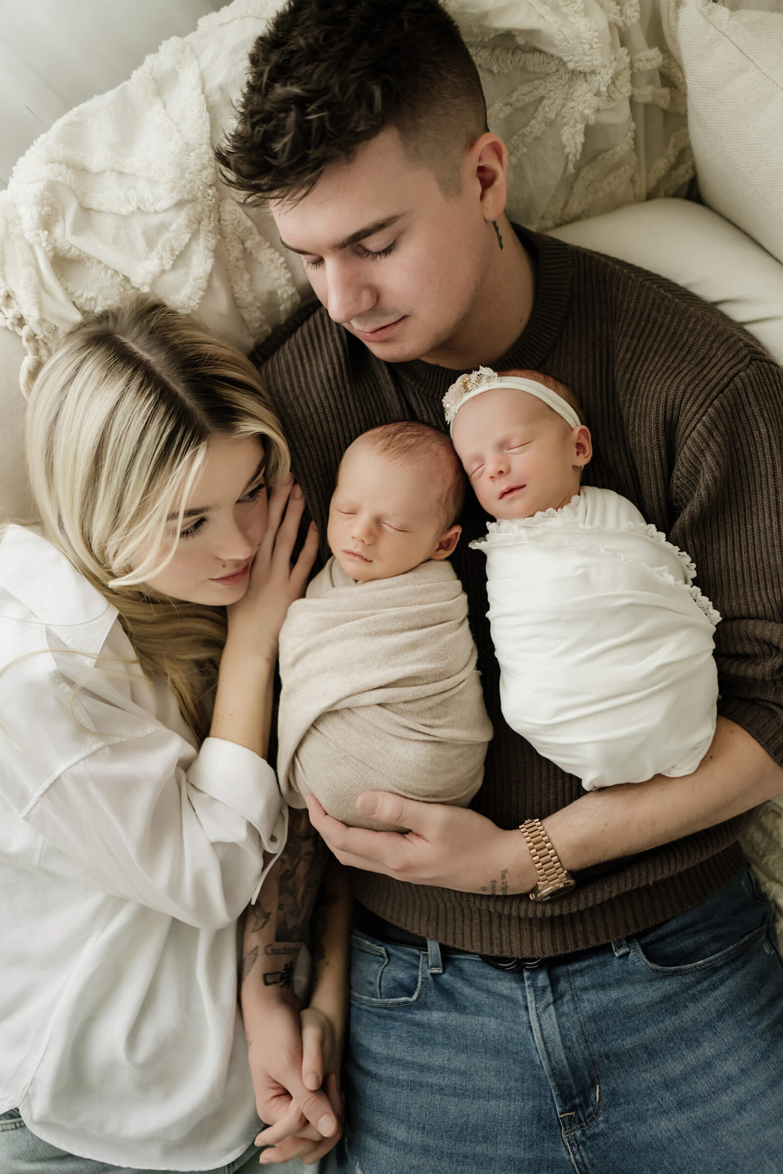 Parents lying together with their newborn twins wrapped in neutral blankets during a calm, connection-centered photo session for newborns.