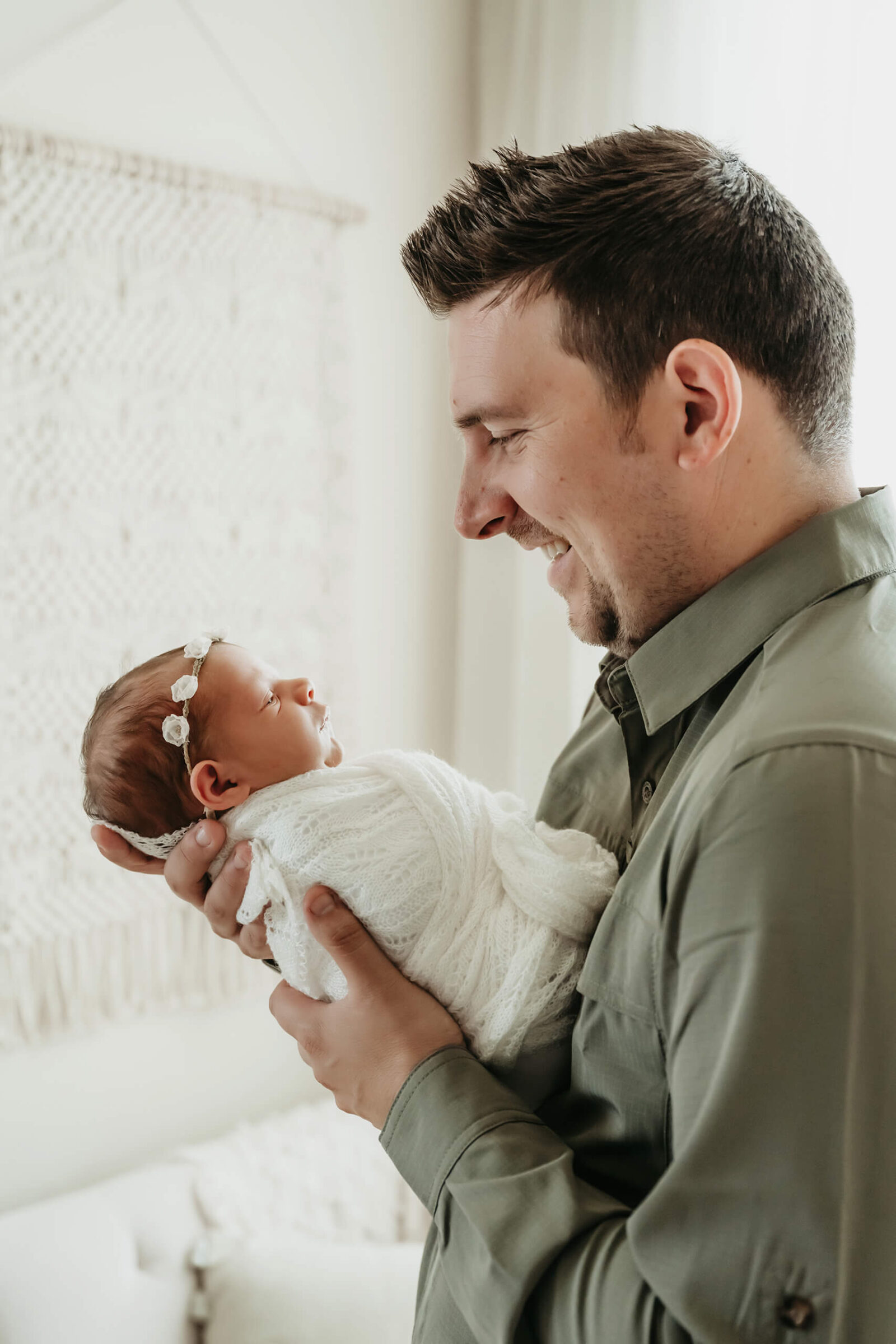 Father smiling while holding his swaddled newborn baby girl during a light-filled, natural studio newborn photoshoot.