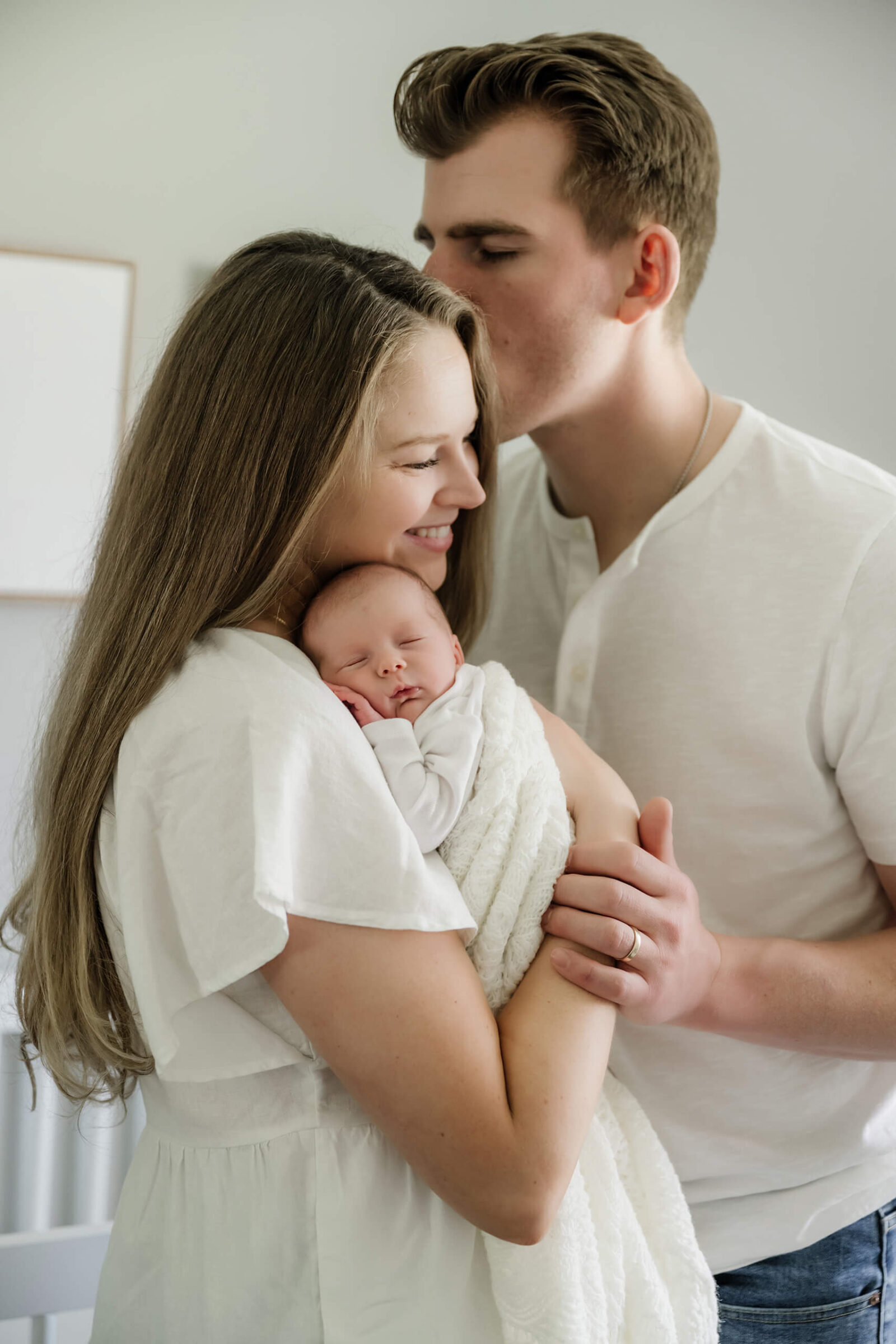 Father kissing mother’s forehead as she holds their sleeping baby in soft window light during a calm and timeless in-home newborn session.