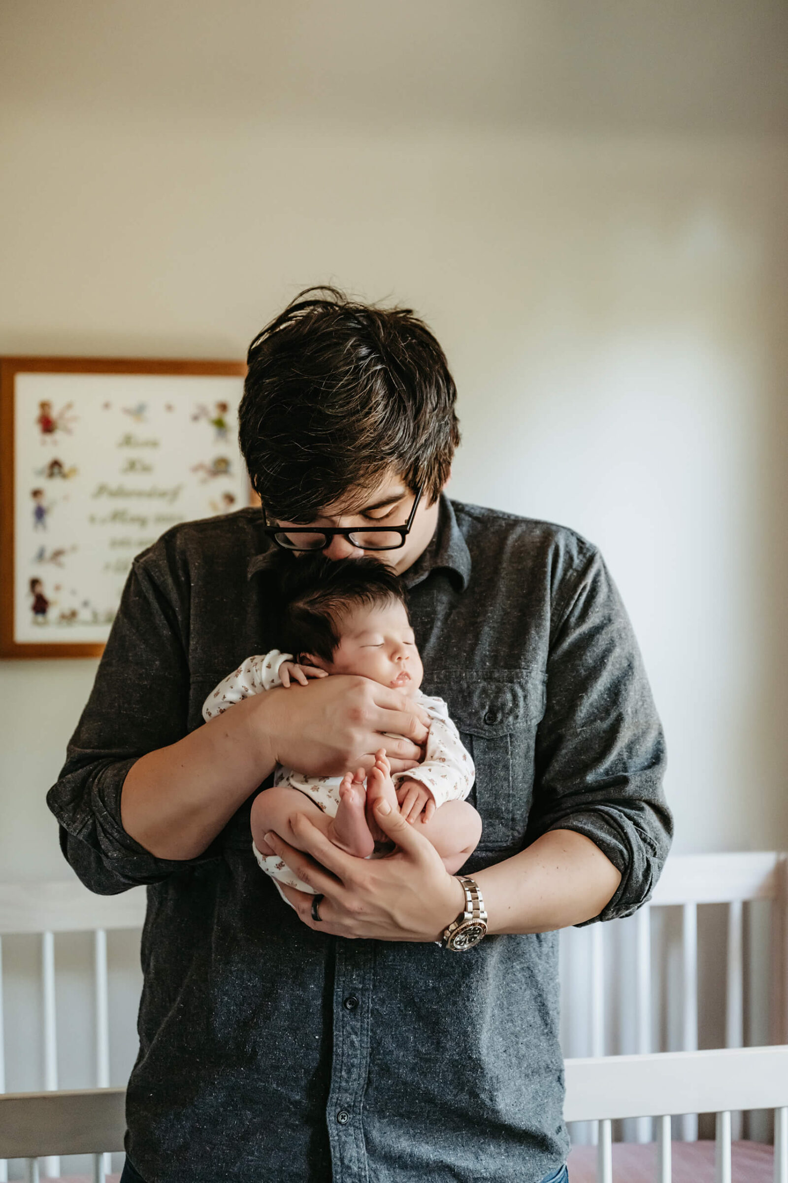 Father cradling his newborn baby close to his chest by the window during a peaceful, light-filled in-home newborn photoshoot.