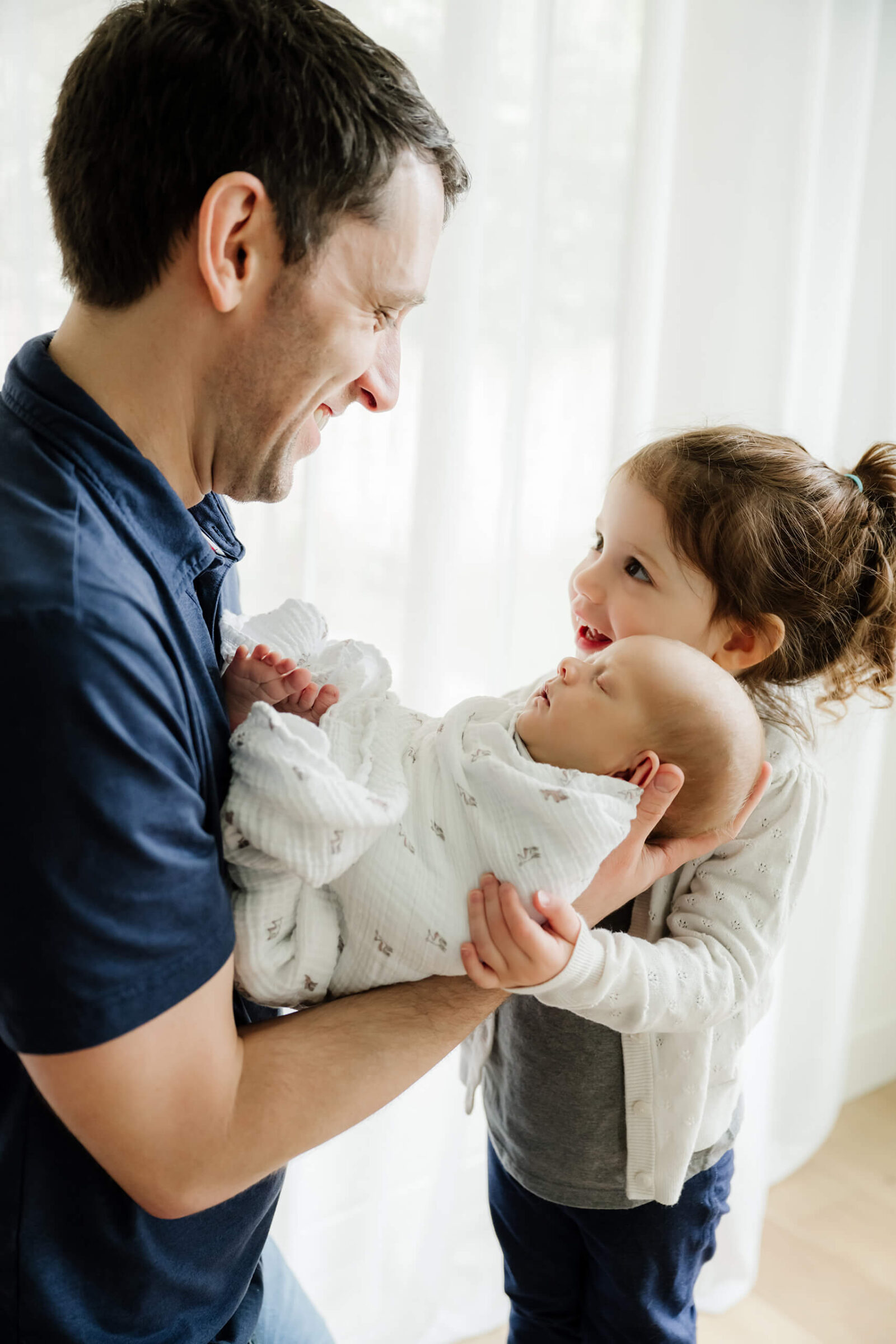 Father and older sister smiling while holding the newborn baby together during a joyful, light-filled in-home newborn photoshoot in Seattle.