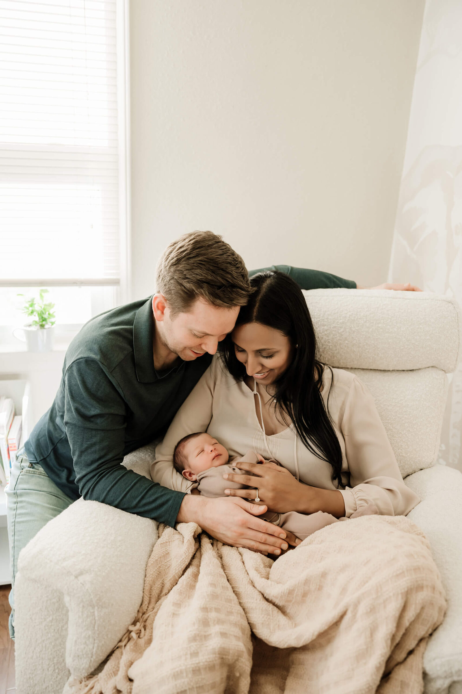 Parents cuddling their newborn baby on a cozy armchair during a warm, natural in-home newborn photo session.