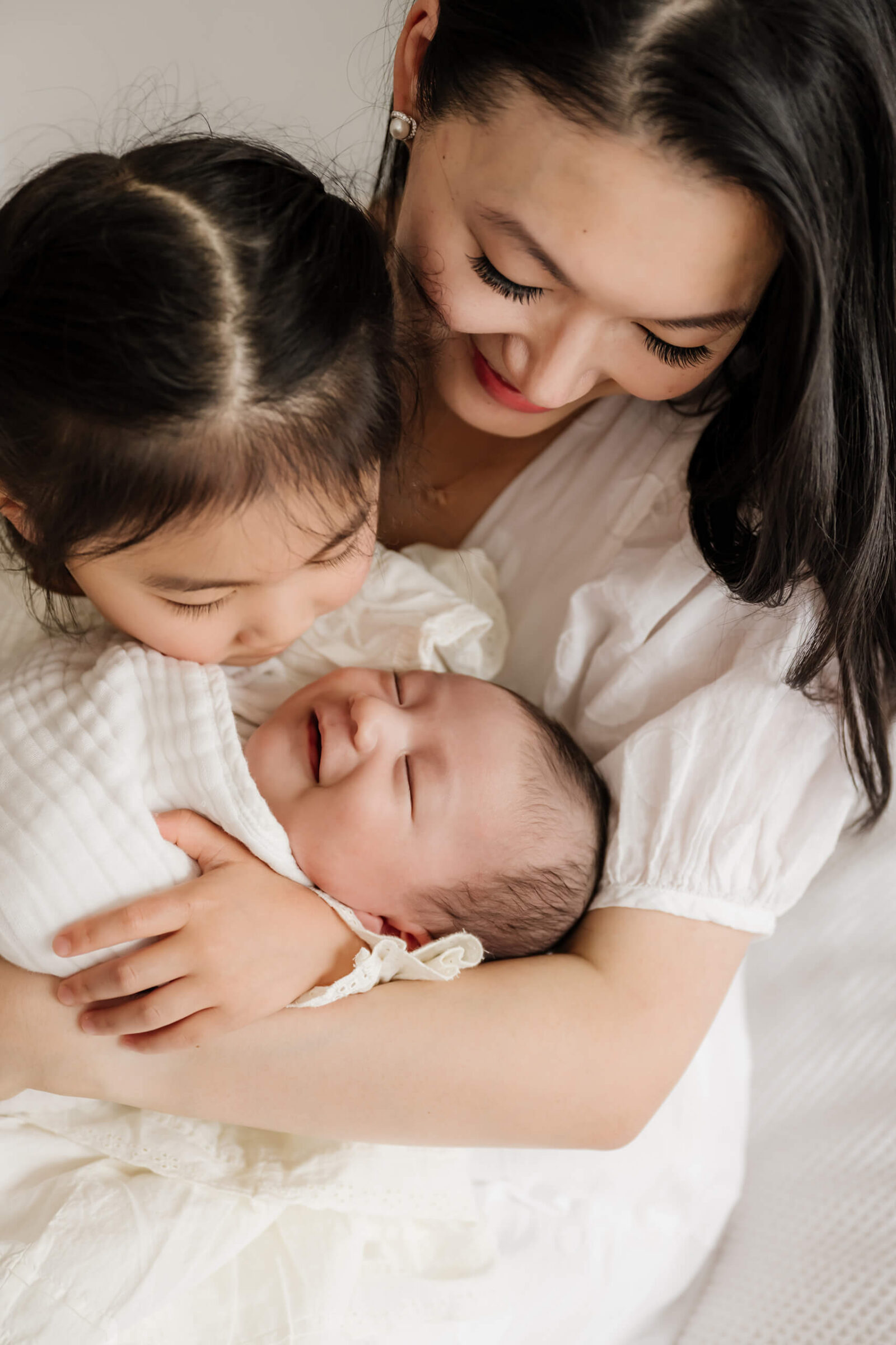 Mother and big sister smiling while snuggling the newborn baby wrapped in white blanket during a tender, love-filled lifestyle newborn session.