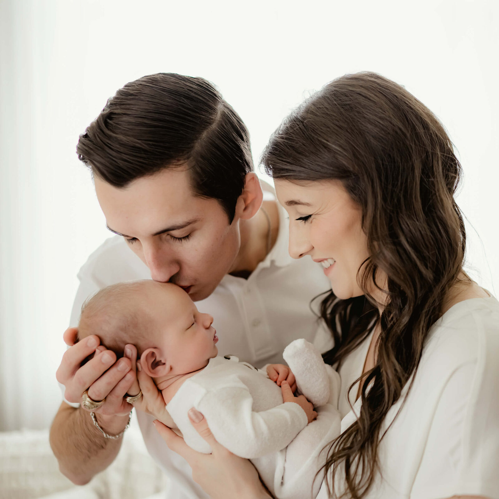 Parents smiling as father kisses newborn’s forehead during a timeless and love-filled photo session for newborn.