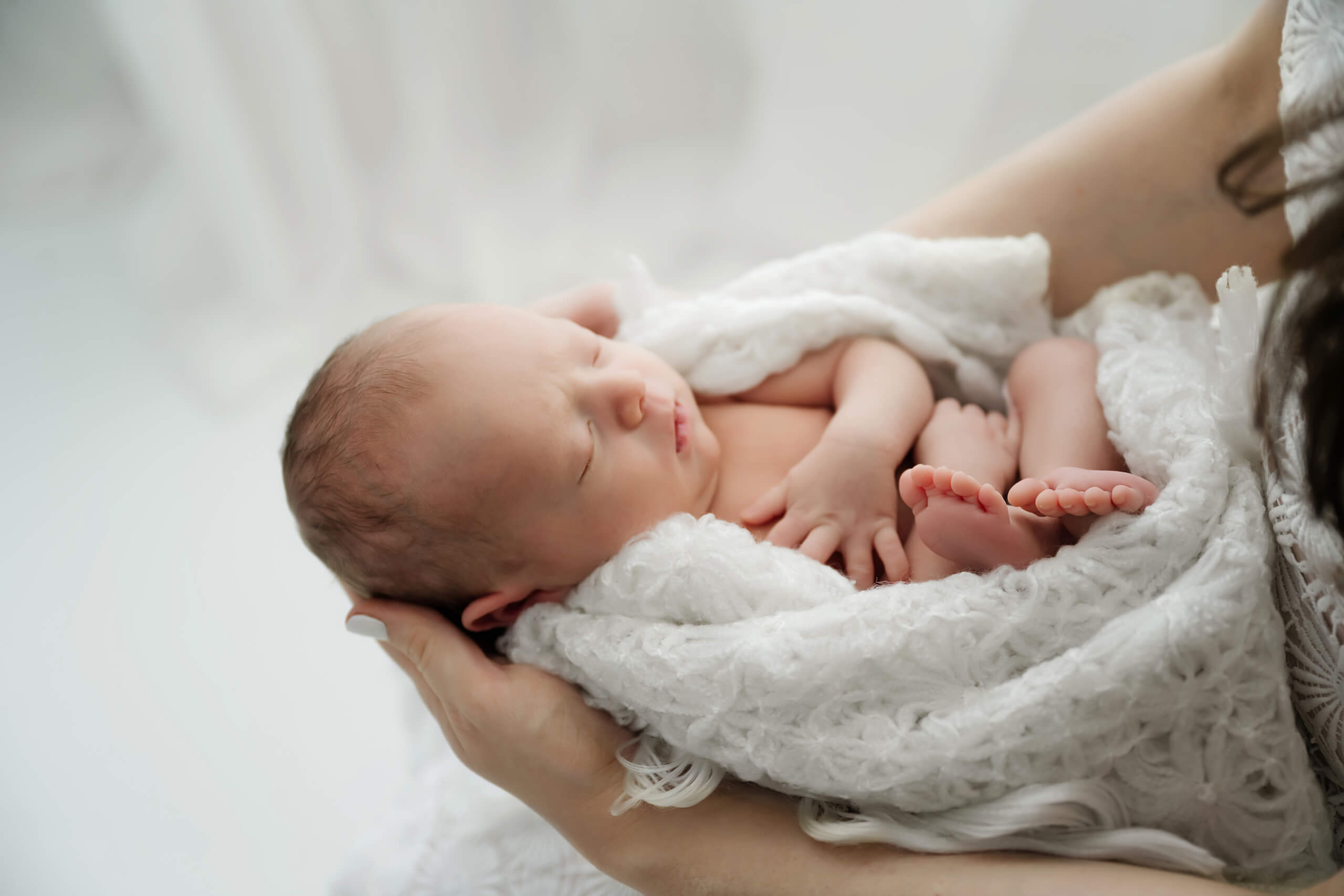 Newborn baby resting in parent&rsquo;s arms wrapped in soft white blanket during a peaceful newborn photoshoot.