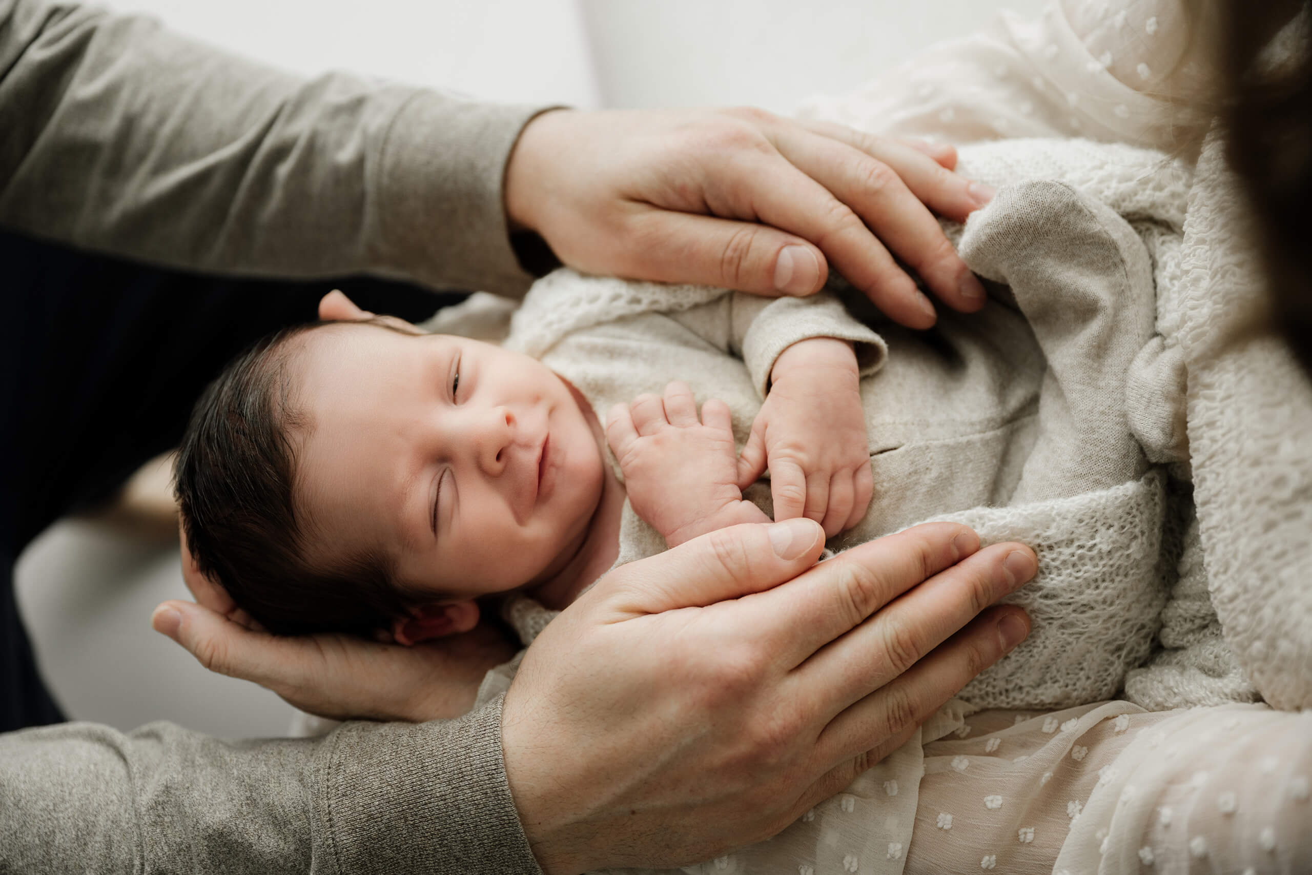Smiling newborn baby surrounded by parents&rsquo; hands during a timeless photo session for newborn.