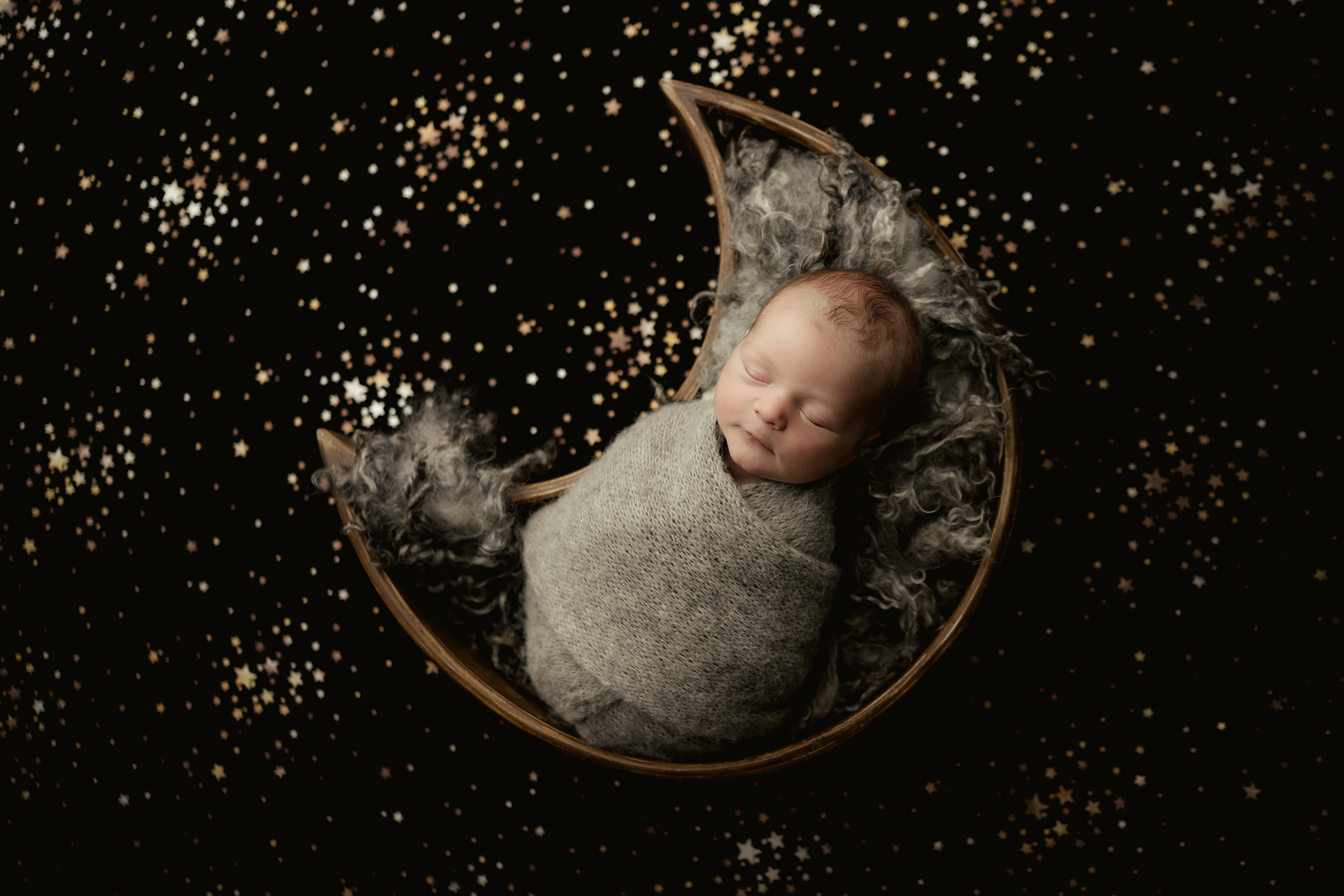 Newborn baby wrapped in gray blanket sleeping in a crescent moon prop during a creative newborn photoshoot.