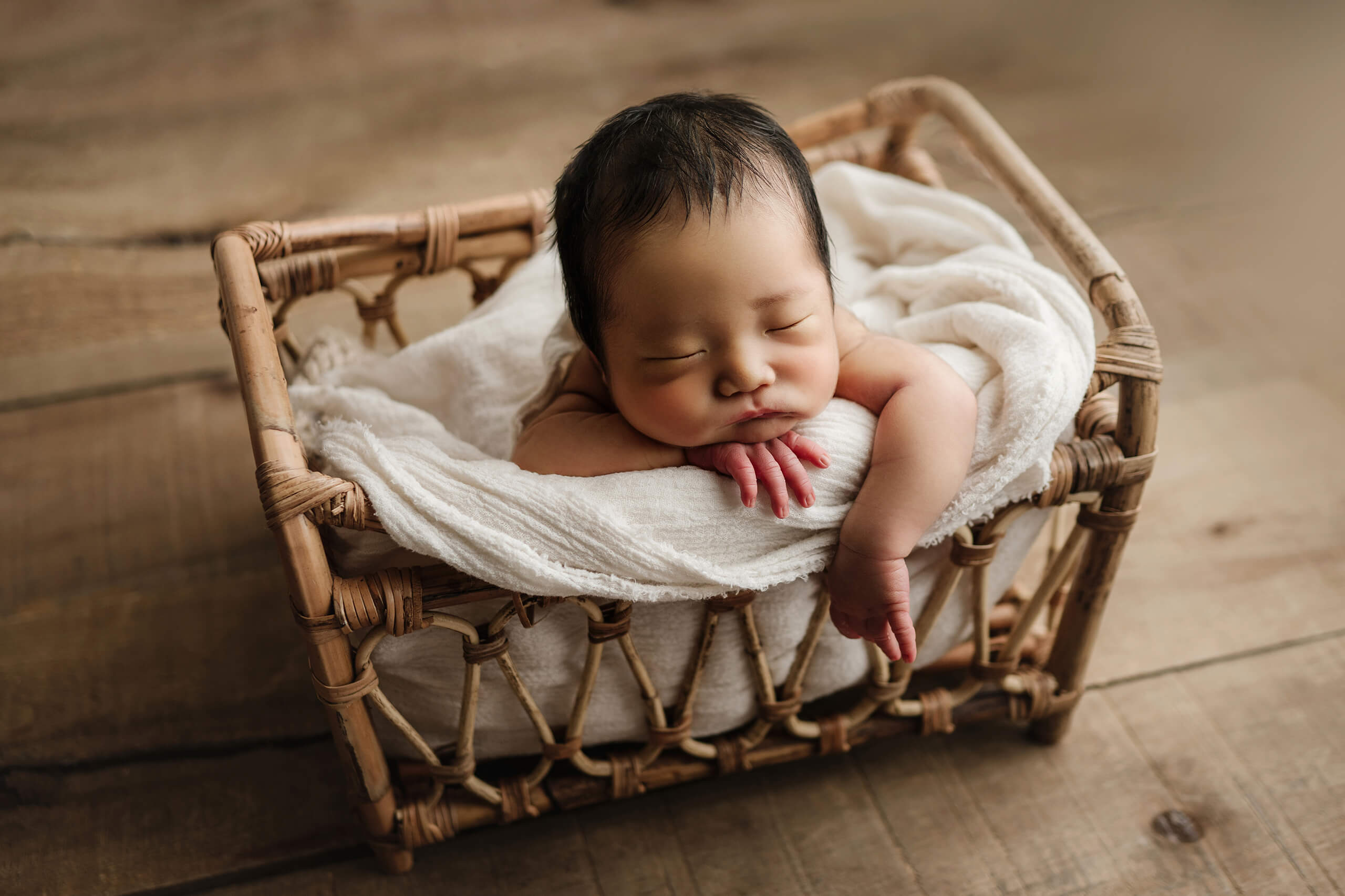 Baby resting in a wicker cradle on wooden floor during a warm, natural newborn photoshoot in Kirkland studio.