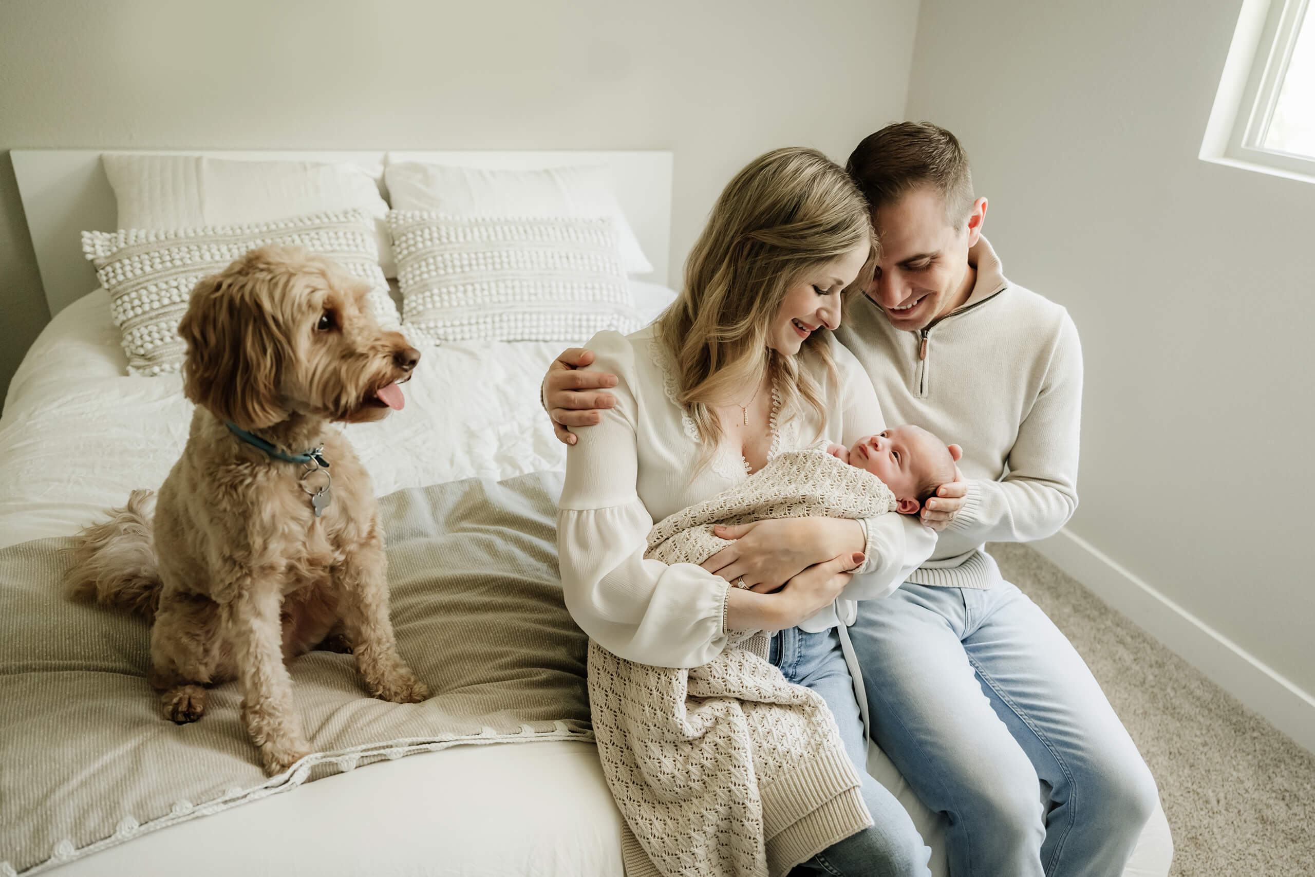Family sitting on a bed with their newborn baby and small dog.