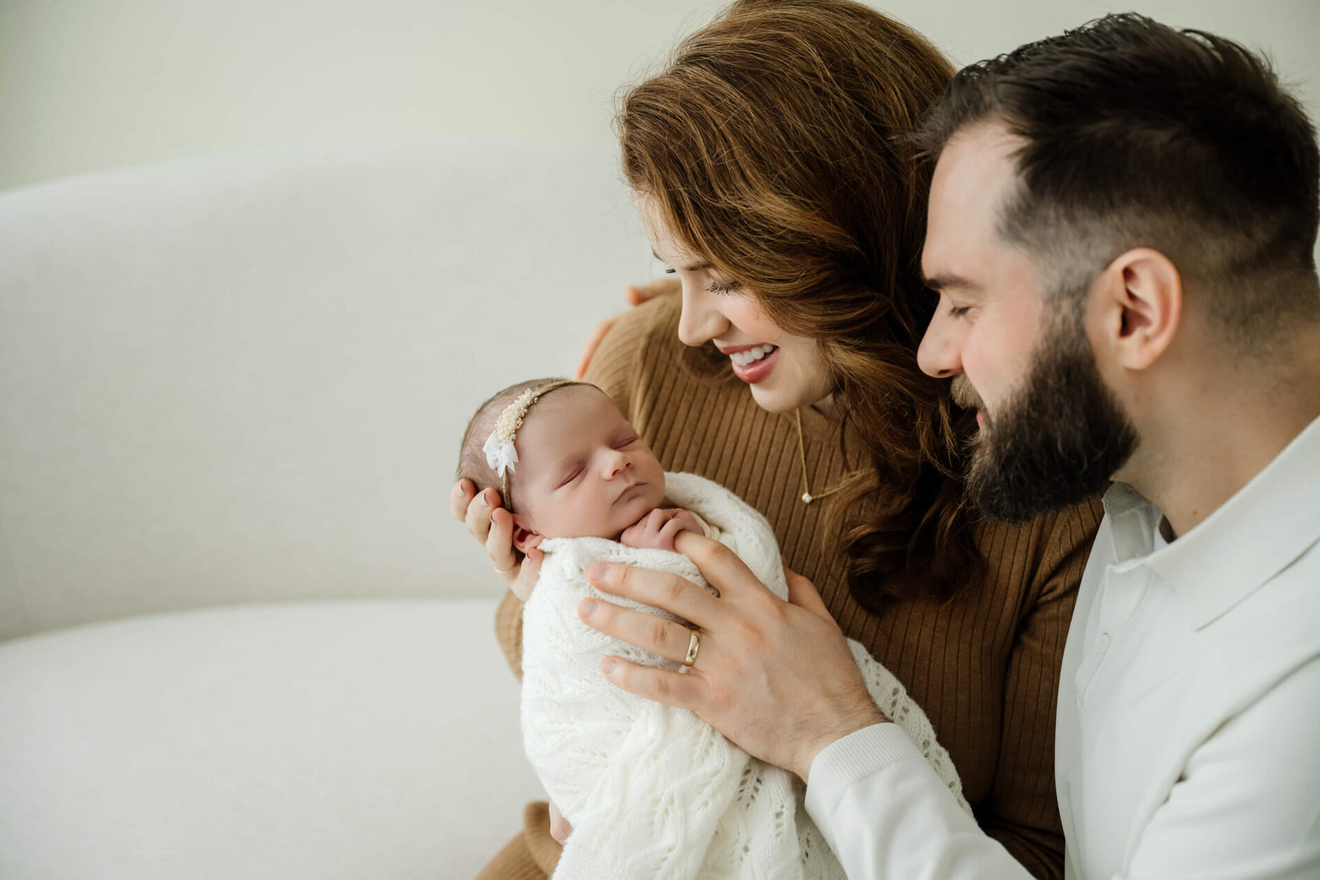 Parents smile down at their swaddled newborn on a studio sofa during a Seattle family newborn photoshoot, cozy neutral styling