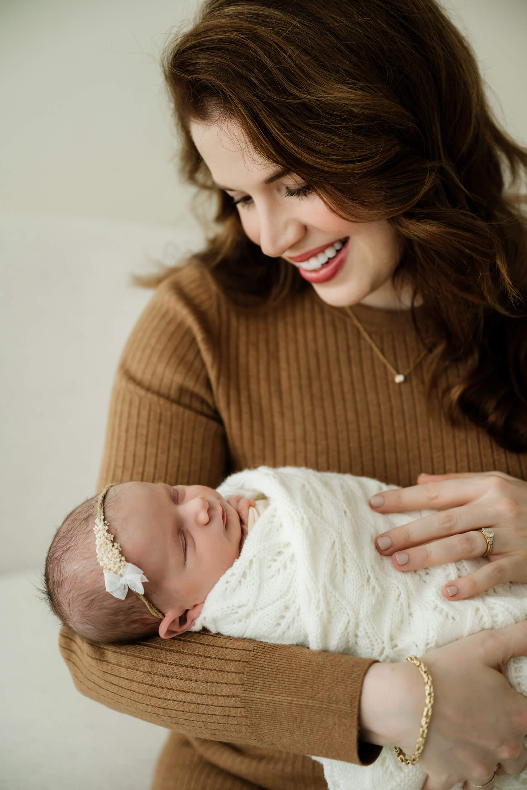 Mother smiles down at her swaddled newborn, warm lifestyle portrait on a neutral backdrop in a Seattle studio
