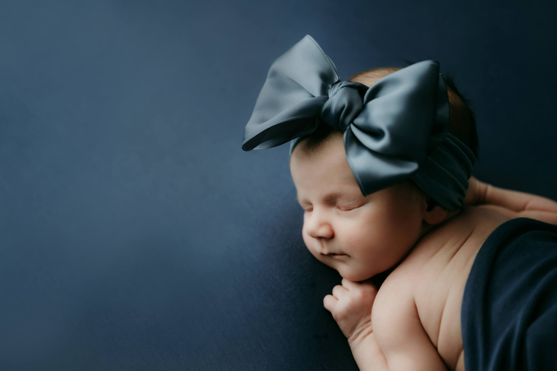 Newborn baby girl sleeps on a deep blue backdrop with a big bow headband in a Seattle studio photoshoot