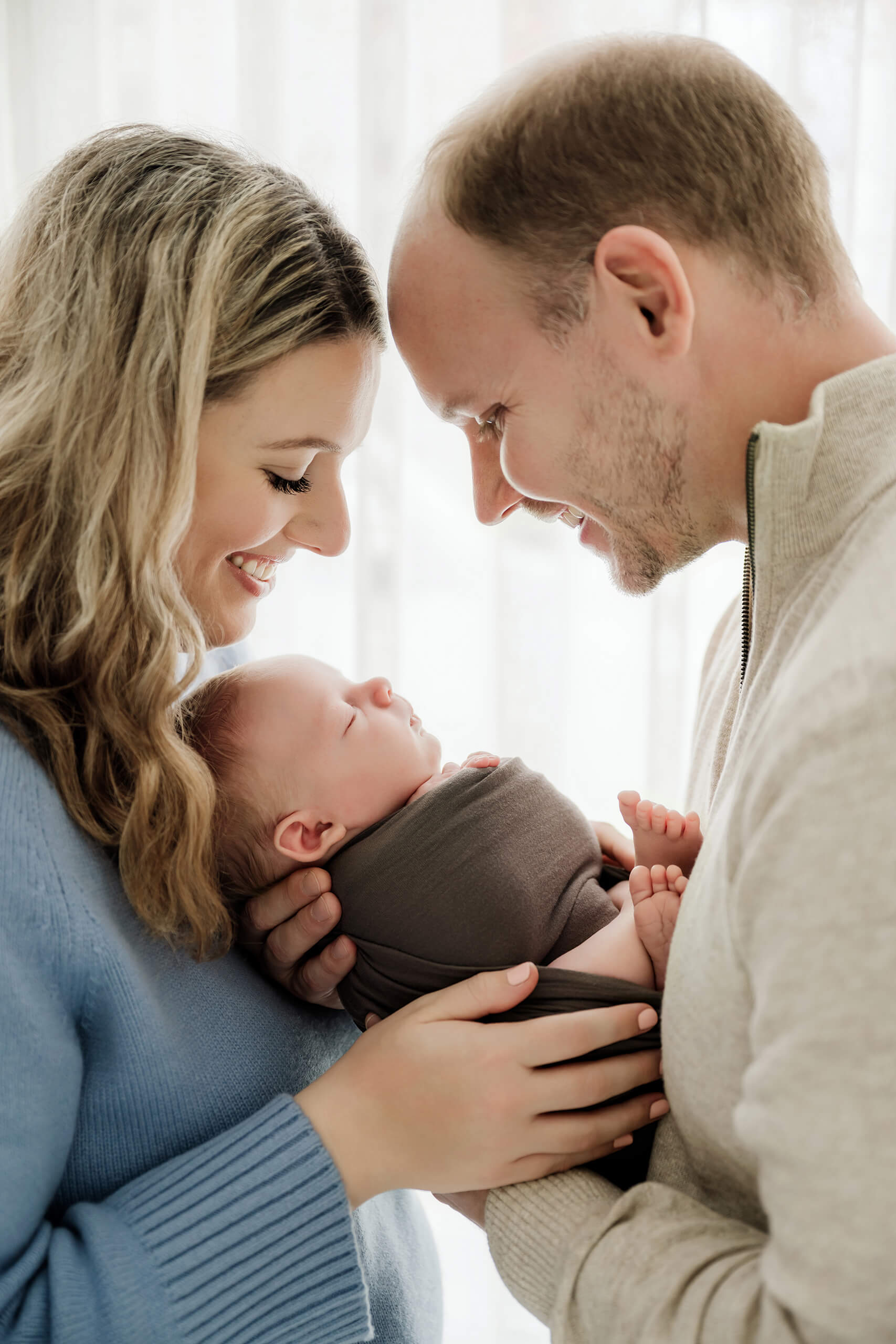 Seattle newborn family photoshoot of parents holding wrapped sleeping baby between them near a bright window.