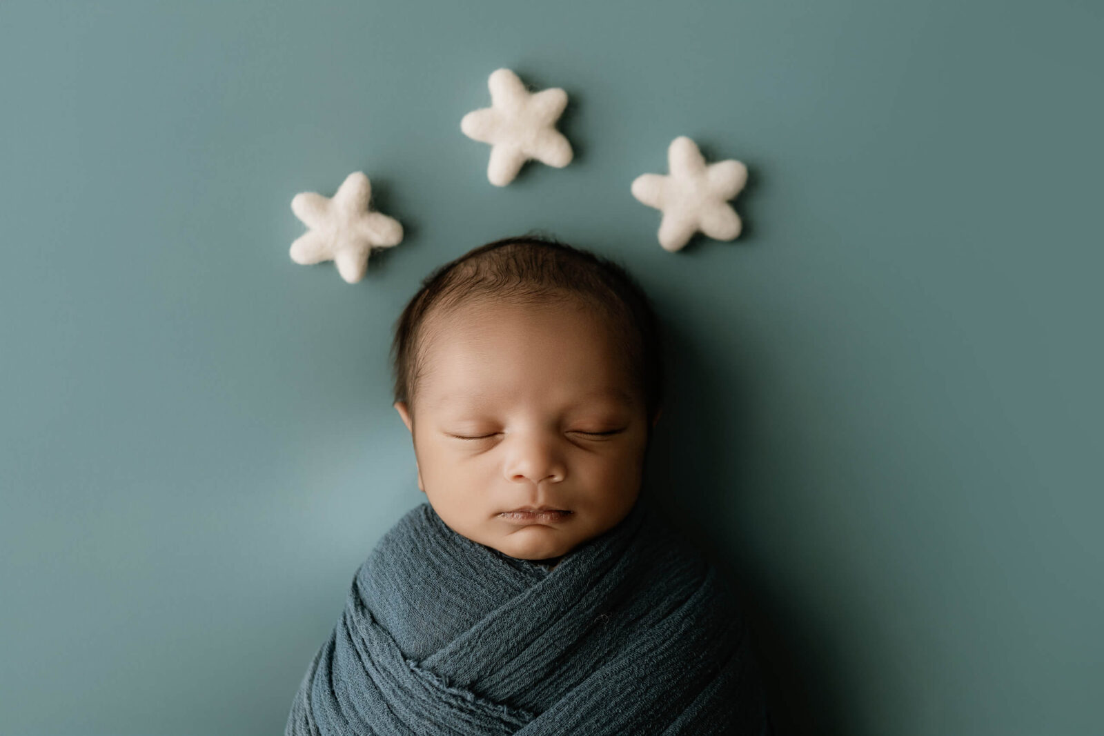 Sleeping newborn wrapped in deep blue fabric on a muted teal backdrop with soft star accents, photographed during a mini studio newborn session in Seattle.