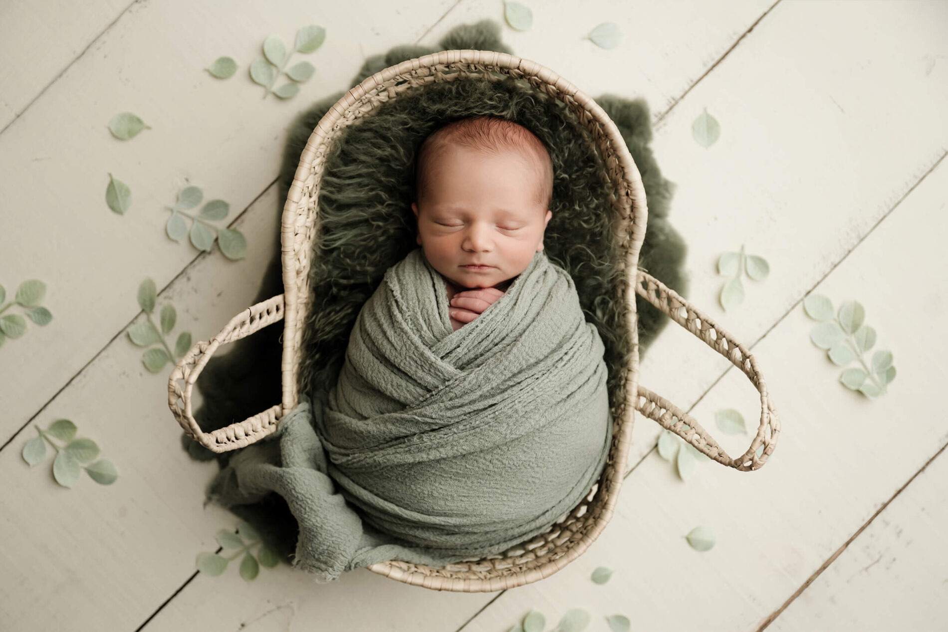 Wrapped newborn sleeping in a woven basket with soft sage tones and delicate greenery, photographed during a mini studio newborn session in Seattle.