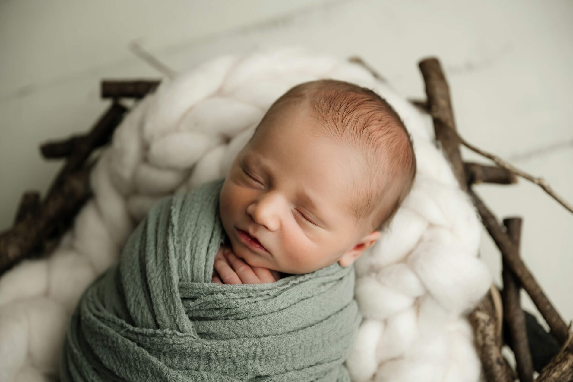 Newborn wrapped in soft green fabric, resting on plush white layers with natural wood accents during a simple studio newborn session in Seattle.
