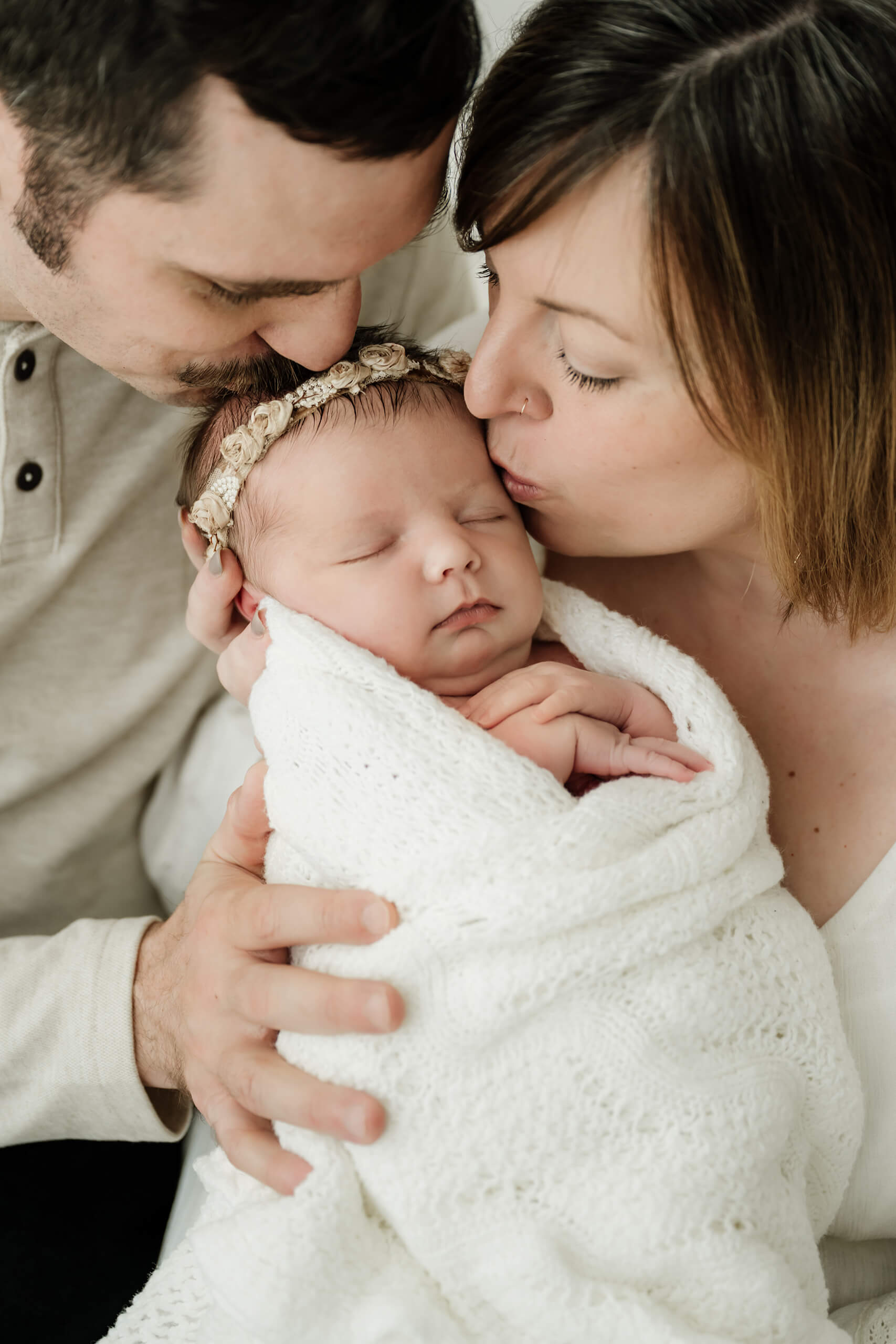 Parents kissing their sleeping newborn wrapped in white, photographed during a simple family newborn session in a Seattle studio.
