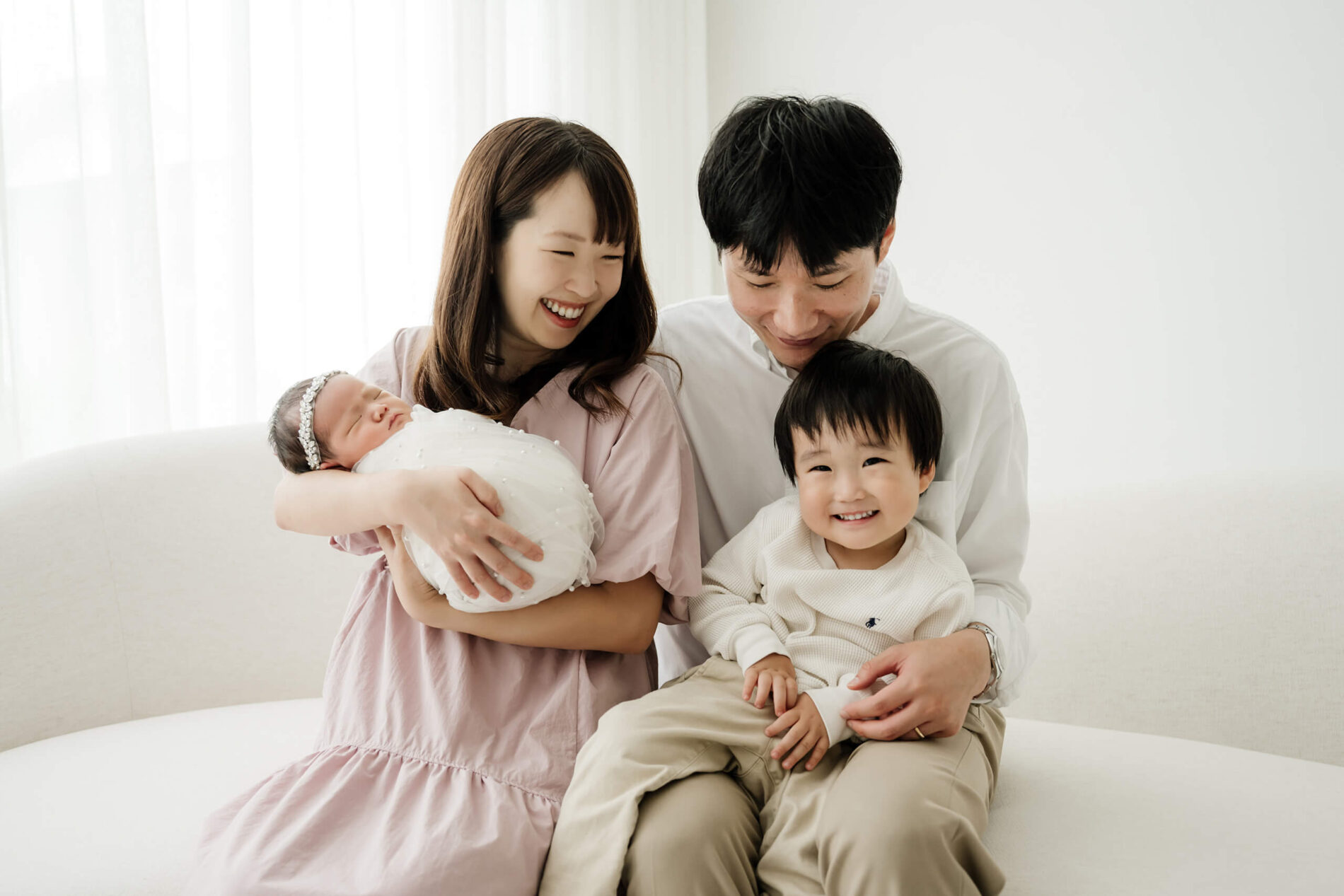 Family sitting together on a studio sofa with parents holding their wrapped newborn and older sibling smiling, photographed during a Seattle mini newborn session.