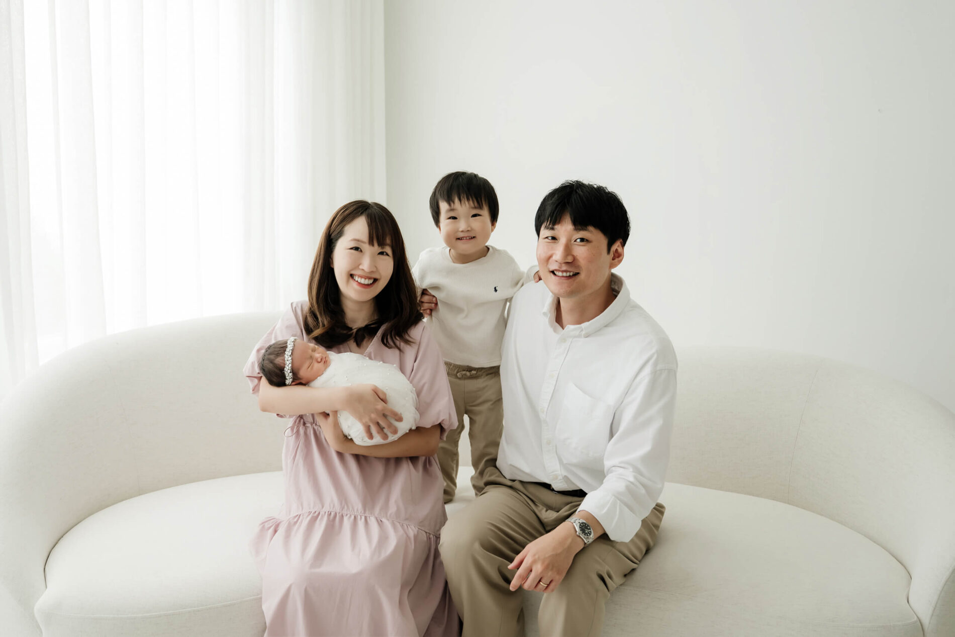 Wide family portrait with parents and toddler sibling gathered around a sleeping wrapped newborn on a curved studio couch in Seattle.