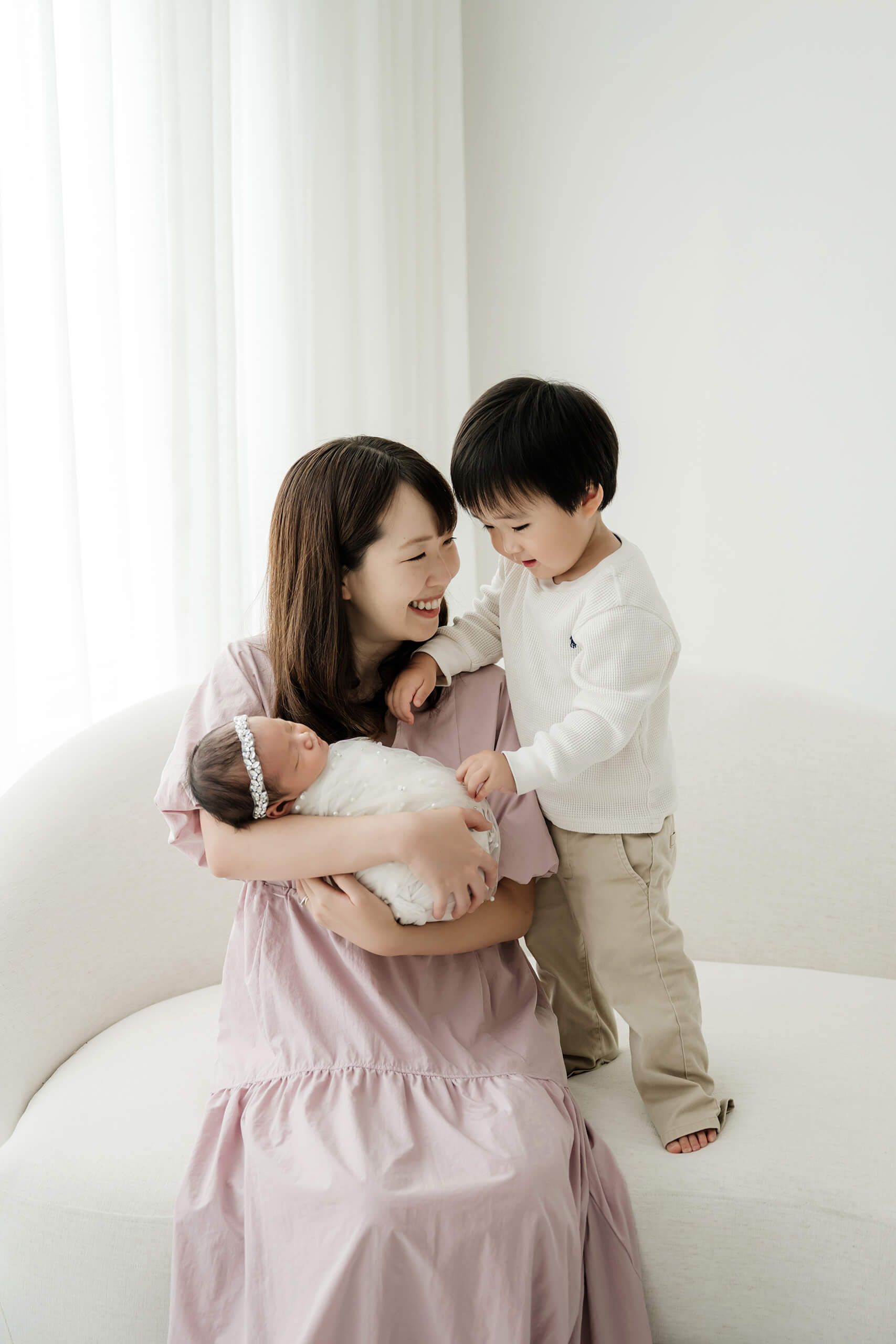Mother seated on a studio sofa holding her newborn while a toddler sibling gently reaches toward the baby, photographed during a family newborn session in Seattle.