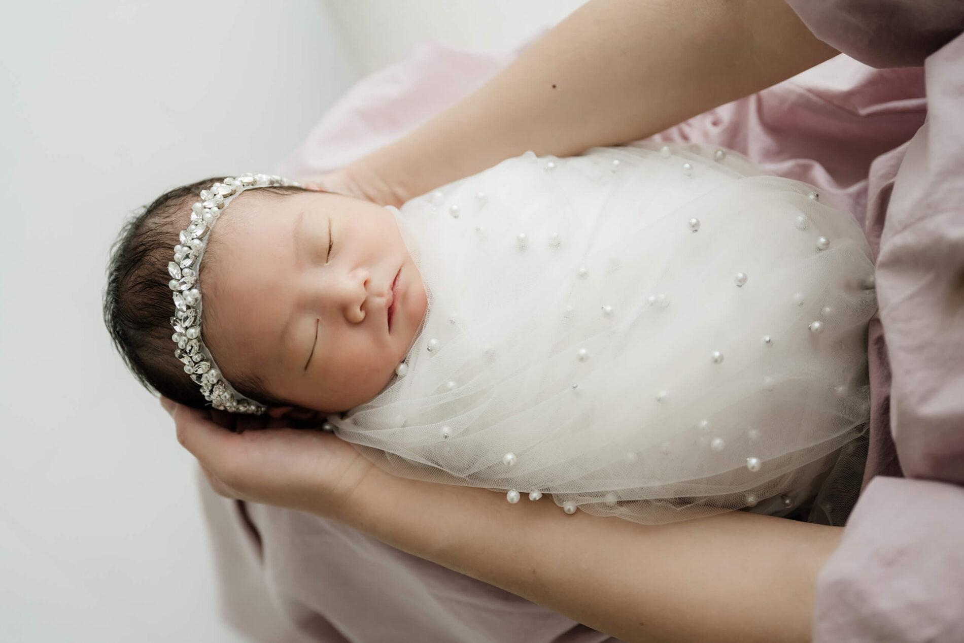 Close-up of a sleeping newborn wrapped in soft white fabric and pearl details, gently cradled in a parent’s arms during a Seattle studio newborn session.