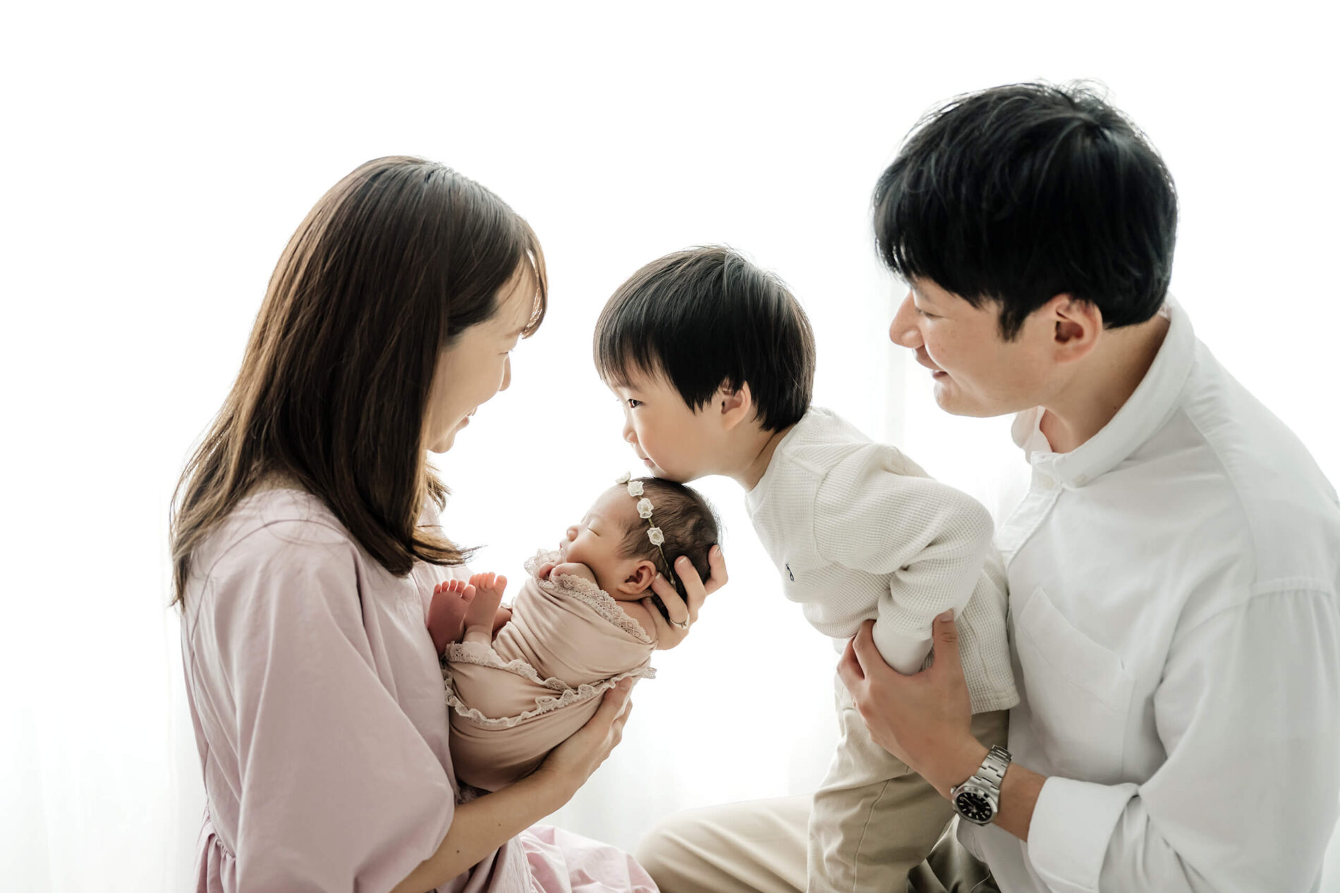 Older sibling leaning in to kiss a sleeping newborn while parents look on, captured in a bright, minimalist Seattle studio newborn session.
